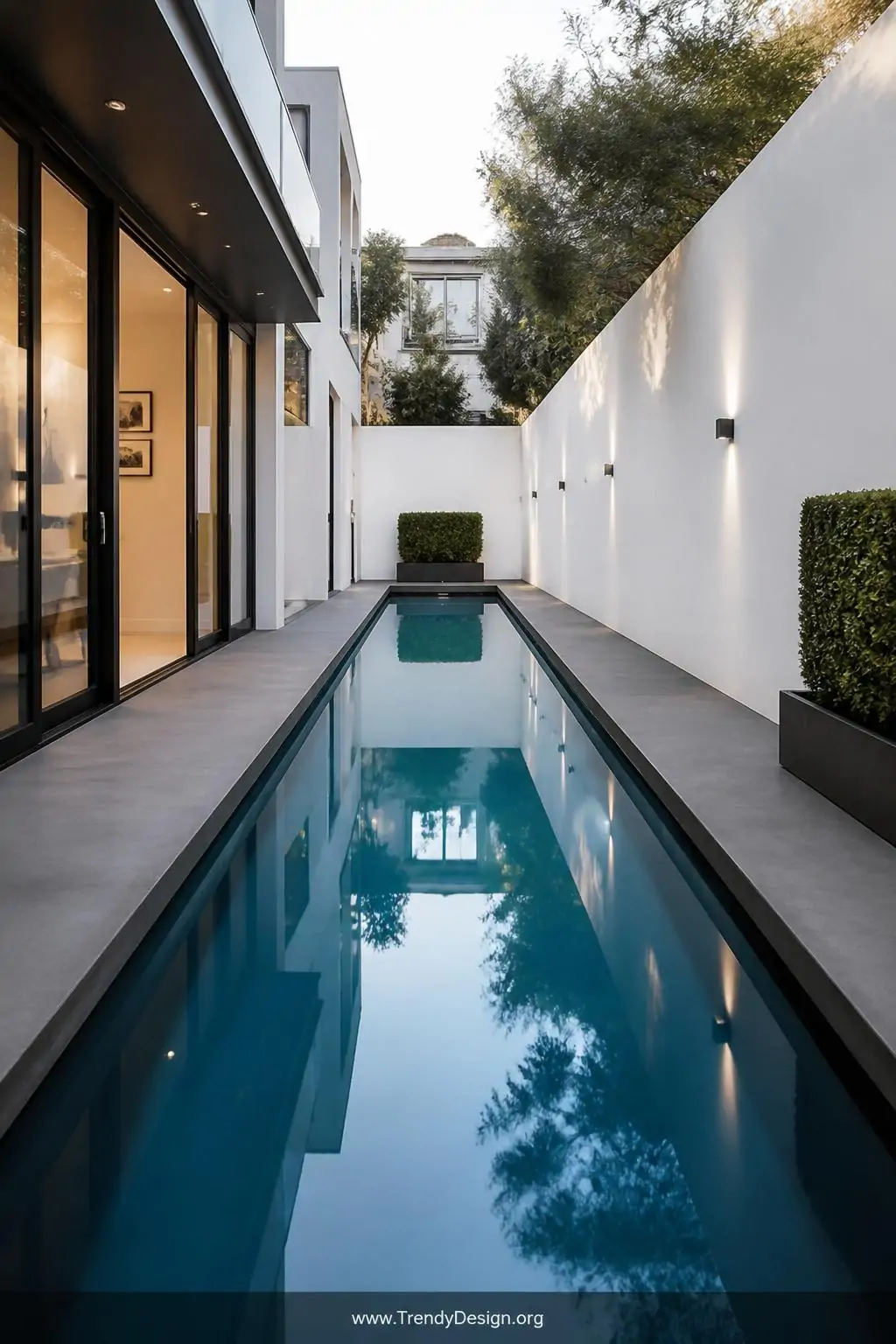 Long narrow lap pool stretching the full length of a sleek modern backyard corridor, charcoal grey concrete deck flanking both sides, crisp white walls reflecting a perfectly still water surface in cool blue tones, minimalist steel planters with clipped boxwood hedges at each end, wide architectural shot, photorealistic interior design.