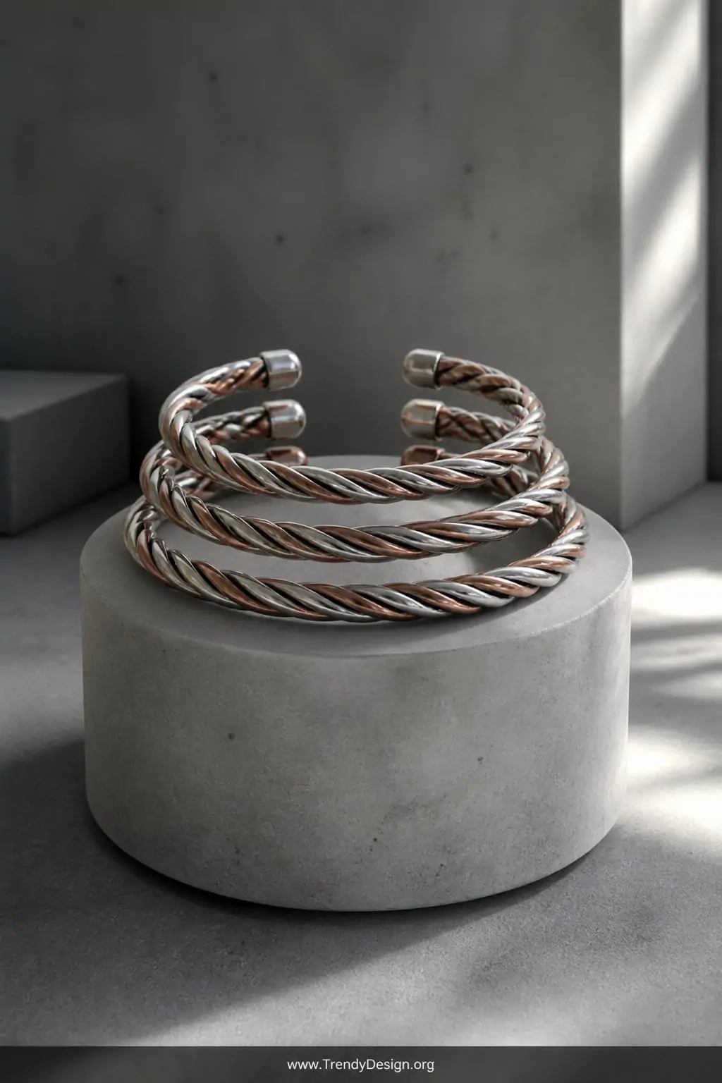 Twisted wire cuff bracelets stacked in a set of three on a brushed concrete jewelry stand, cool studio lighting, copper and silver two-tone rope-texture bands arranged in ascending size order, medium shot