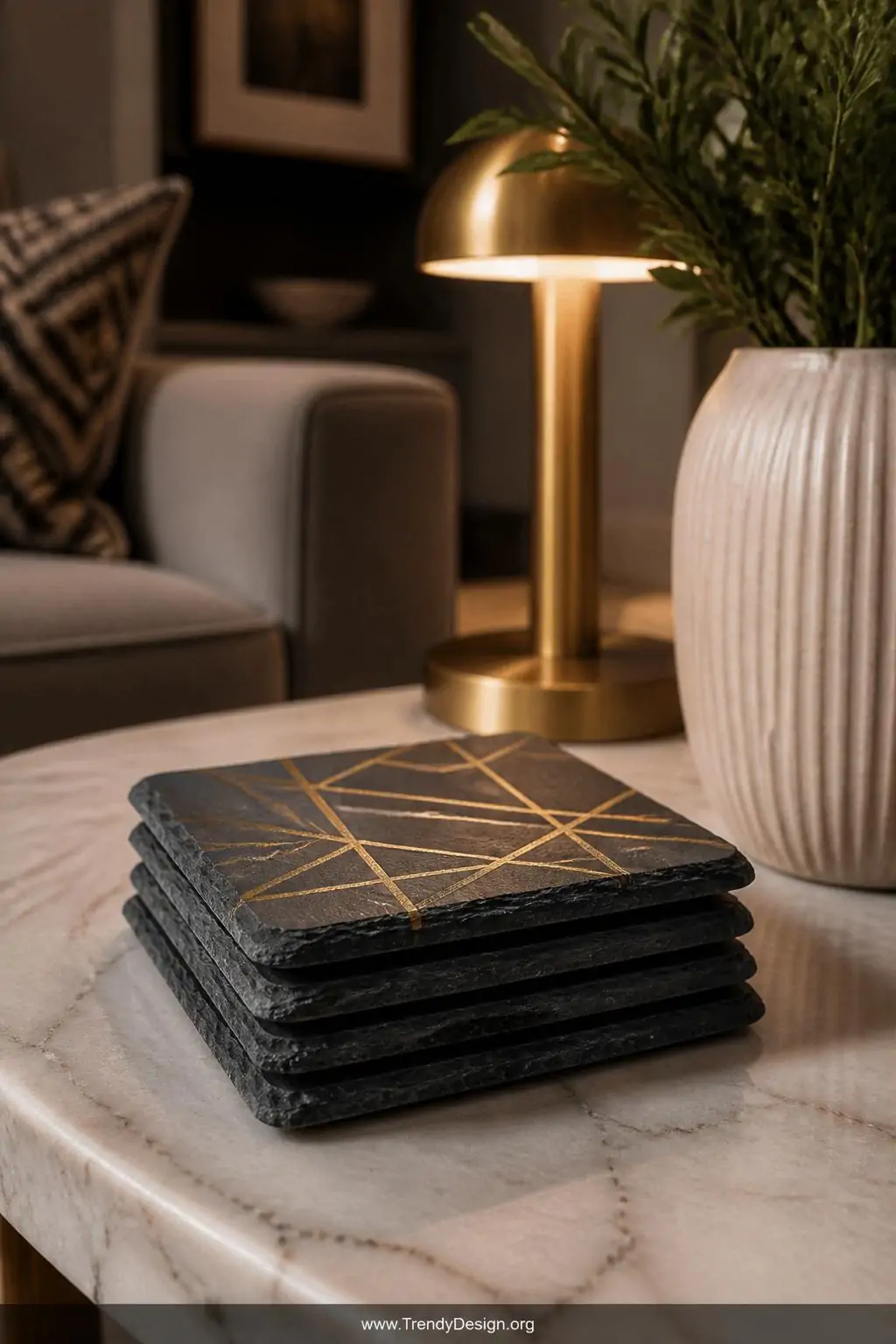 Set of four square dark slate coasters with gold metallic geometric painted designs and felt-padded bottoms stacked on a marble coffee table, modern eclectic living room, soft warm side lamp light, closeup shot, photorealistic interior design, no people.