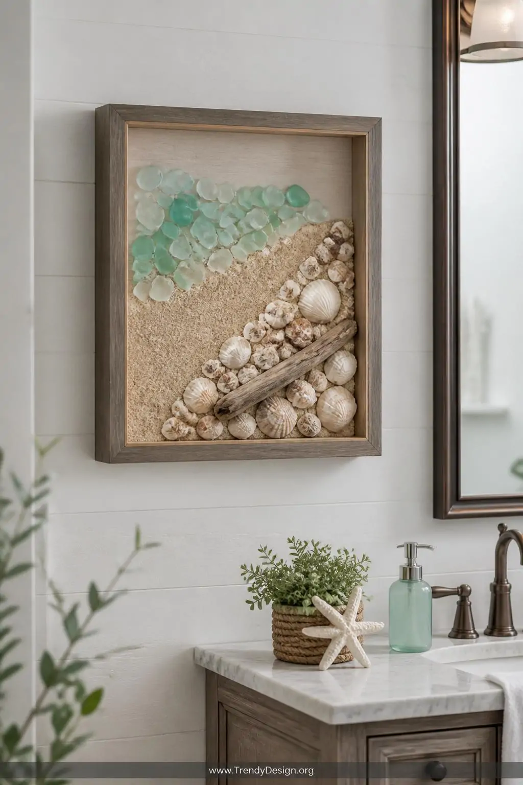 Deep shadow box frame mounted on a whitewashed bathroom wall displaying a sand-coated base with arranged sea glass, clustered shells, and a small driftwood piece forming a beach scene, soft diffused light, medium shot