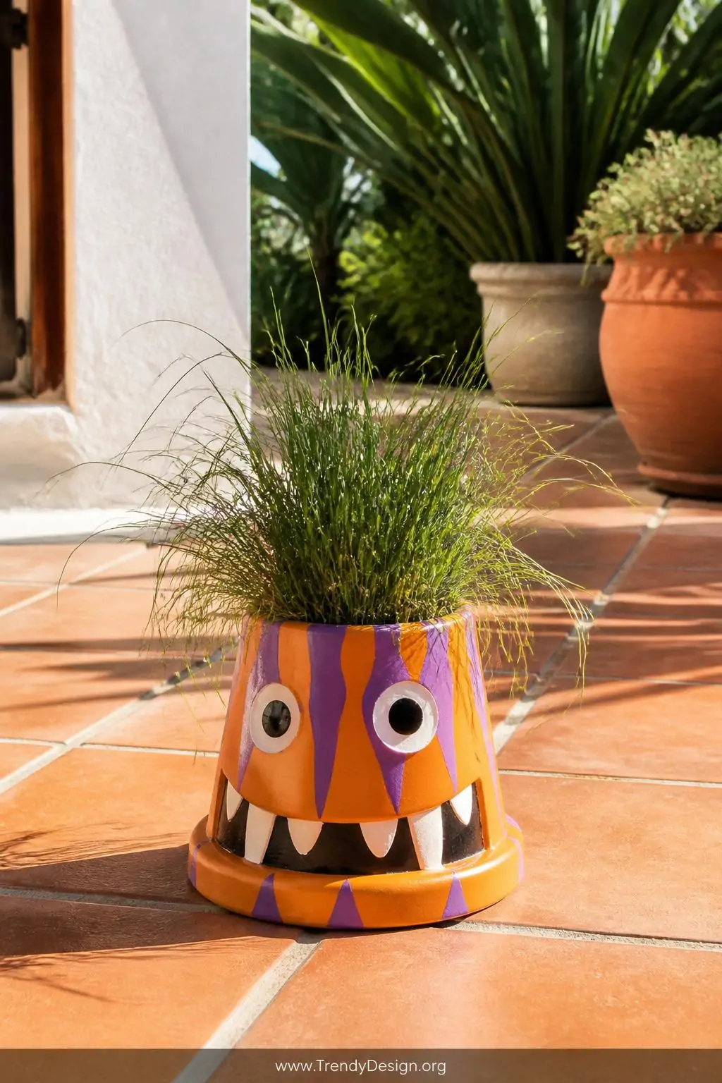 Upside-down terracotta pot painted with wild orange and purple monster fangs and googly eyes sitting on a sun-drenched terracotta tile patio surface, bright outdoor afternoon light, thick wild green grass erupting dramatically from the drainage hole on top, medium shot.