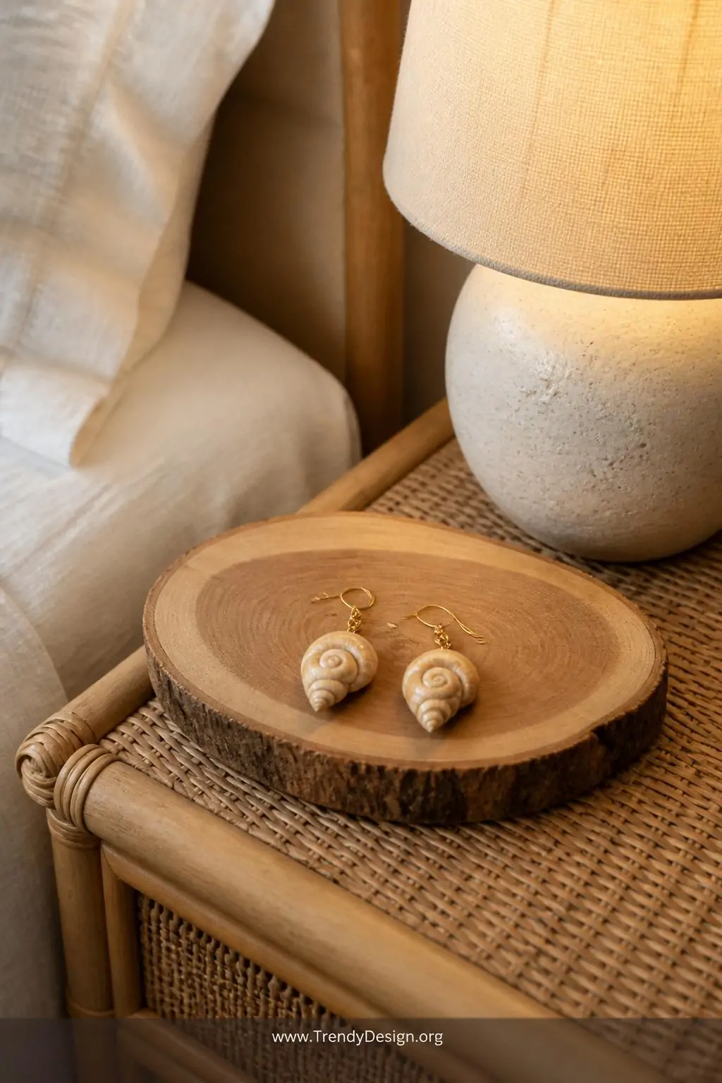Pair of spiral auger shell earrings on gold fish hook wires resting on a raw-edge cedar jewelry dish atop a rattan nightstand, soft warm bedside lamp glow, cream linen textures in background, closeup shot, photorealistic interior design
