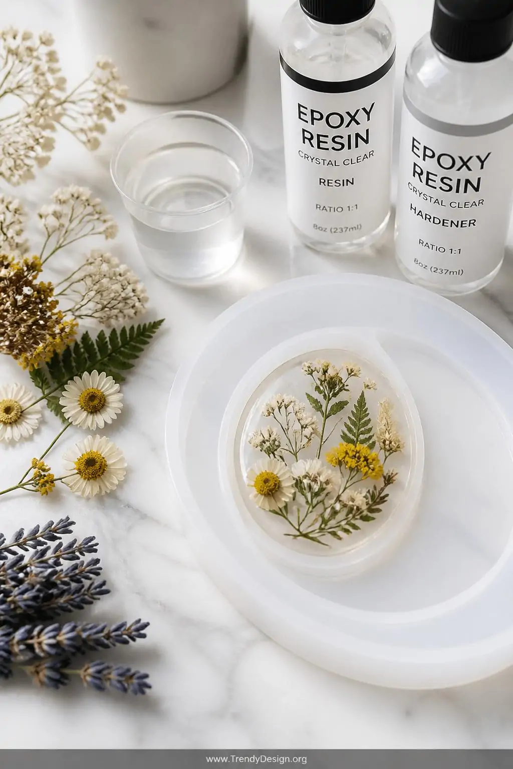 Clear resin brooch with suspended pressed wildflowers and botanicals curing in a silicone mold beside dried florals and a two-part resin kit on a marble white craft countertop, bright natural daylight, closeup