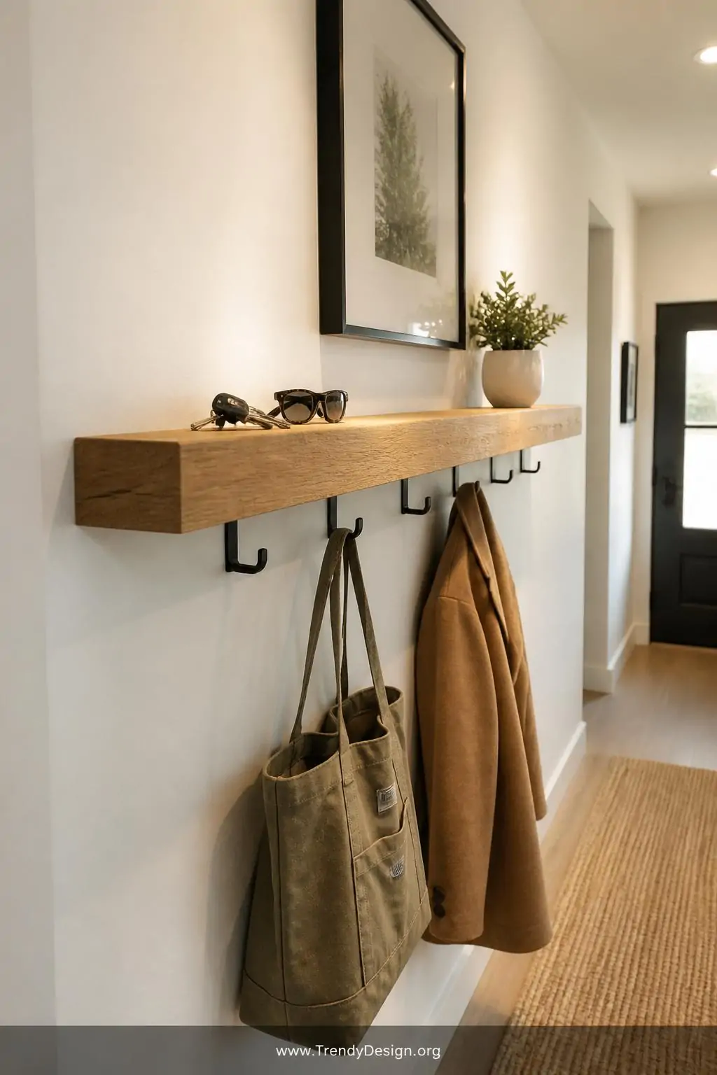 Chunky oak floating shelf with matte black industrial hooks underneath mounted in a narrow white entryway, keys and sunglasses resting on the top surface, olive canvas tote and camel wool coat hanging below, warm recessed lighting highlighting the golden wood grain against crisp white walls, medium shot, photorealistic interior design.