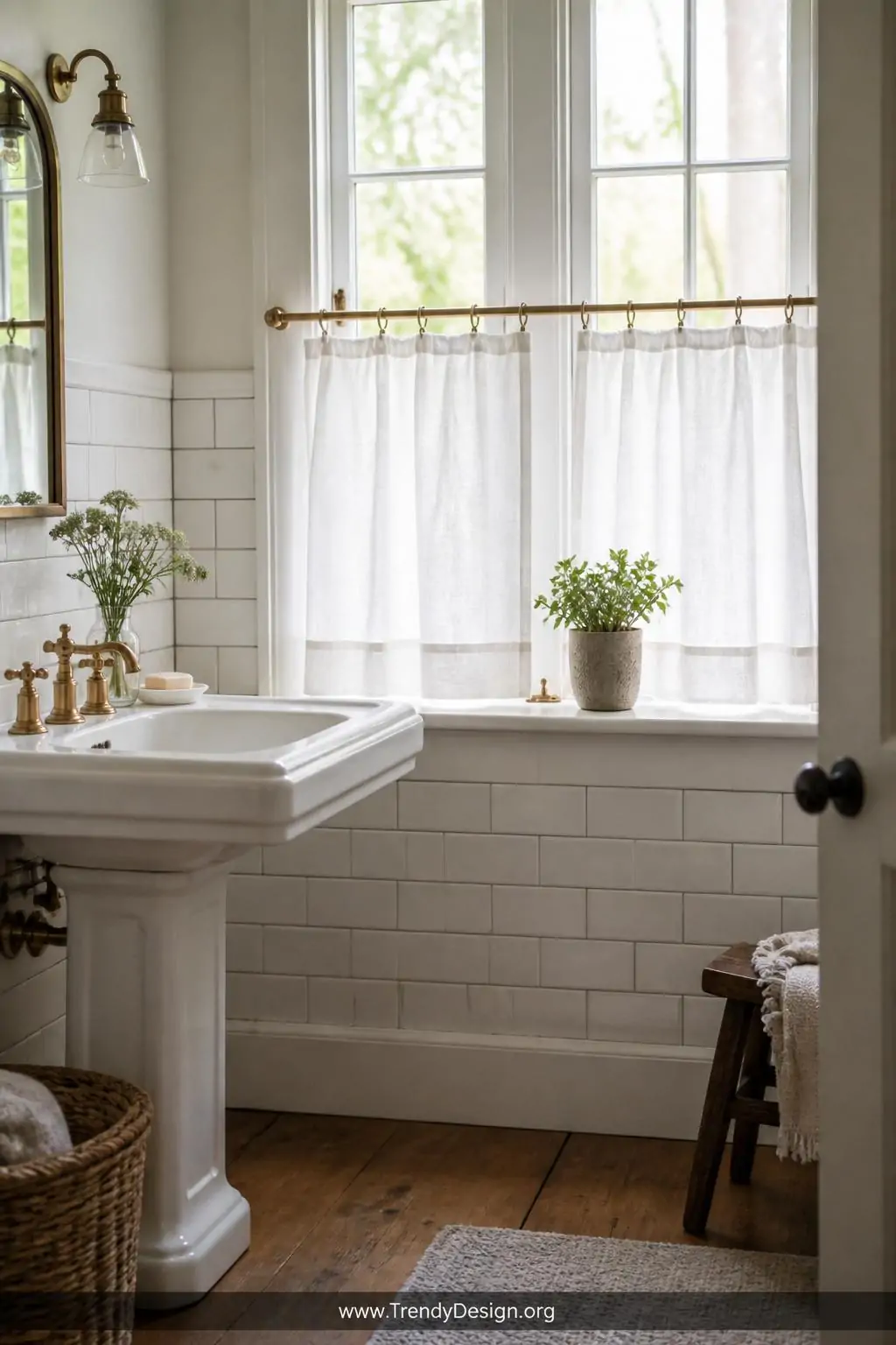 Medium shot of a charming farmhouse bathroom showcasing crisp white linen café curtains hung at mid-window on a slender brass