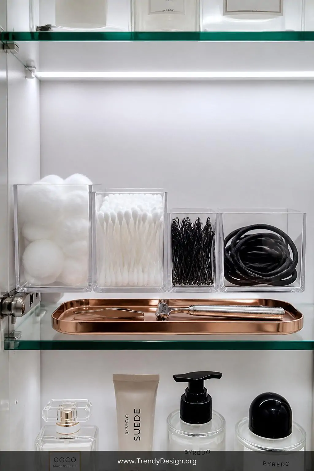 A tight closeup of a single glass medicine cabinet shelf lined with clear acrylic bins holding cotton balls, cotton swabs, bo
