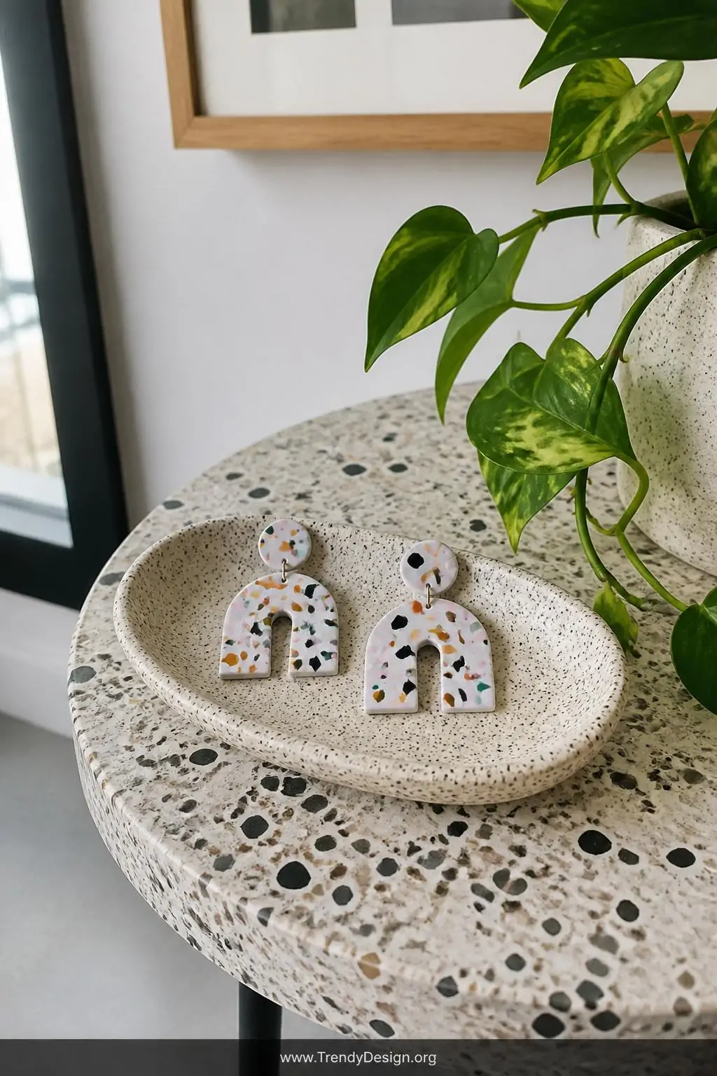 Abstract terrazzo polymer clay earrings with colorful confetti speckle surfaces on a white and grey base displayed on a speckled stone catchall dish beside a trailing pothos plant on a terrazzo-topped side table, bright natural daylight, medium shot showing the playful designer surface detail.