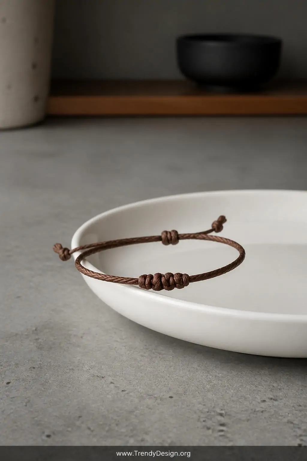 Minimalist single-strand waxed linen sliding knot bracelet draped over a smooth white ceramic dish on a sleek concrete countertop, soft diffused overhead lighting, tight closeup revealing the precise knot structure against a cool minimalist Japandi kitchen shelf aesthetic.