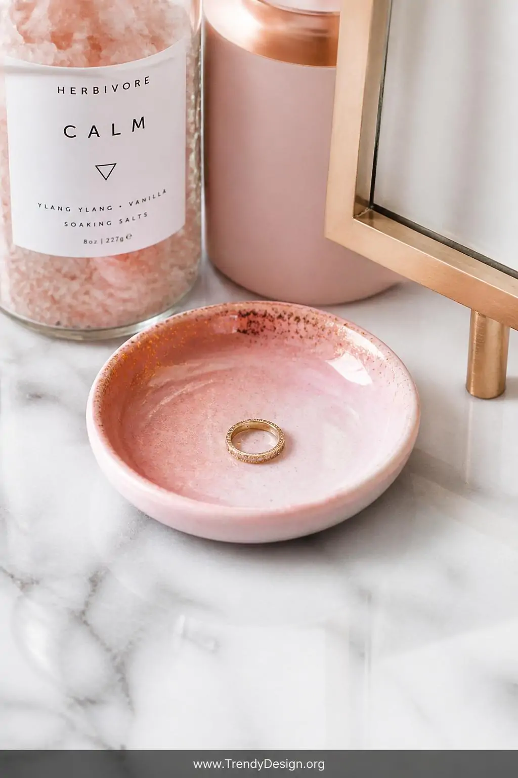 Petite dip-dyed clay ring dish in blush ombre with rose gold interior shimmer holding a delicate gold ring on a white marble vanity beside bath salts, soft diffused light, closeup shot