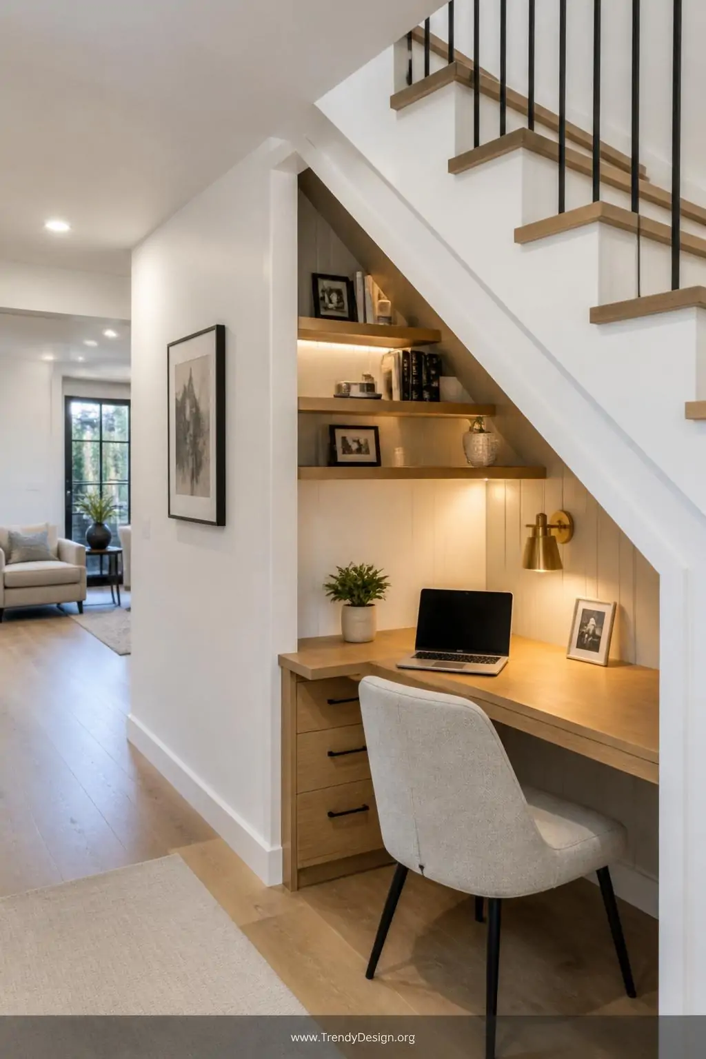 Custom triangular under-stair desk built flush into an angled white wall, warm wood desktop with wall-mounted shelves following the descending staircase slope overhead, a small brass sconce casting focused warm light across the workspace, wide shot of an open-plan hallway home office nook, contemporary minimalist style, photorealistic interior design.