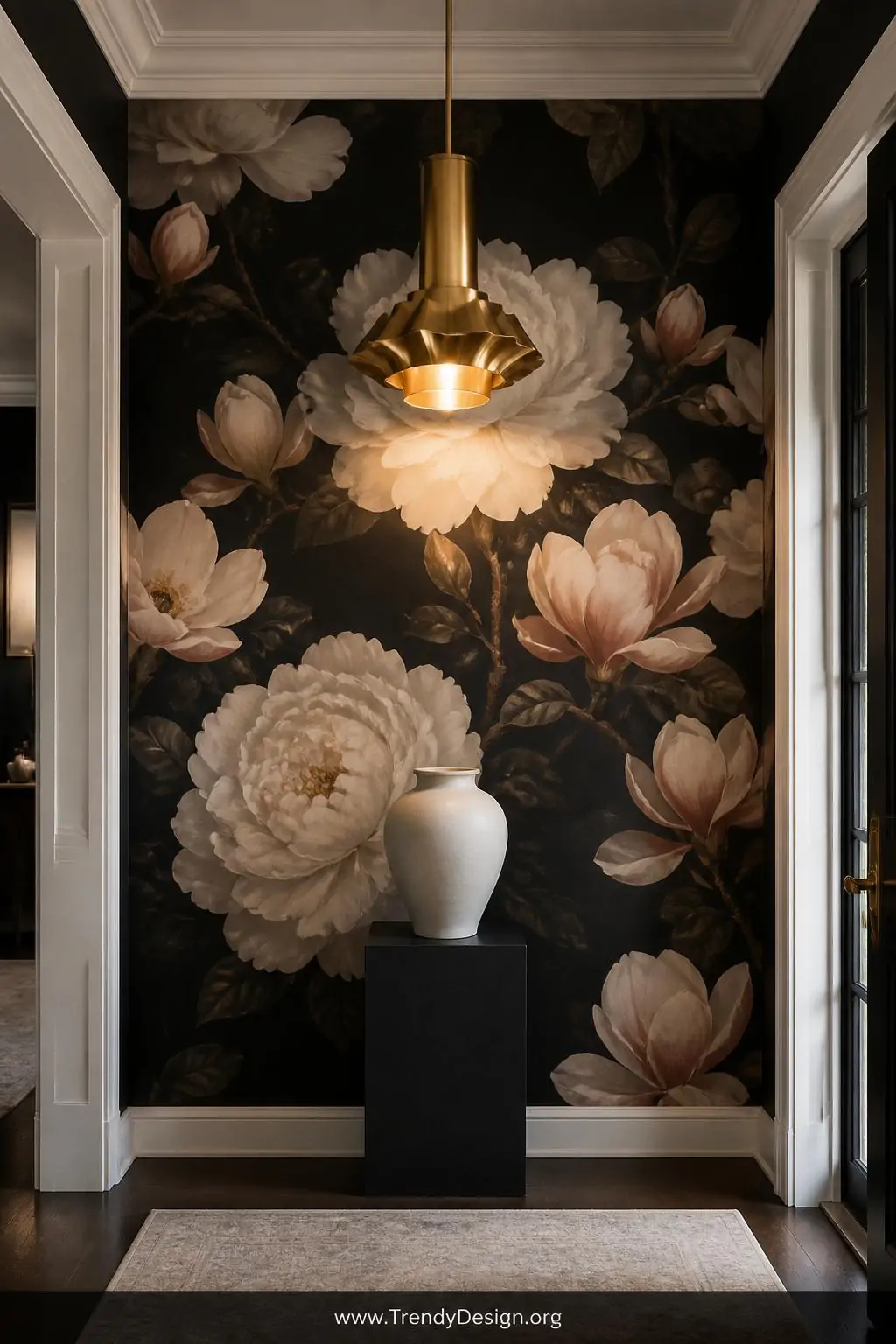 Moody dark floral wallpaper with oversized ivory peonies and blush magnolias blooming against a deep charcoal near-black background in an entrance hallway, dramatic contemporary style — a brushed gold statement pendant light hangs centrally casting warm amber glow across the blooms, a single cream ceramic vase sits on a dark plinth below, crisp white ceiling and woodwork framing the darkness, medium shot centered on the glowing floral wall.