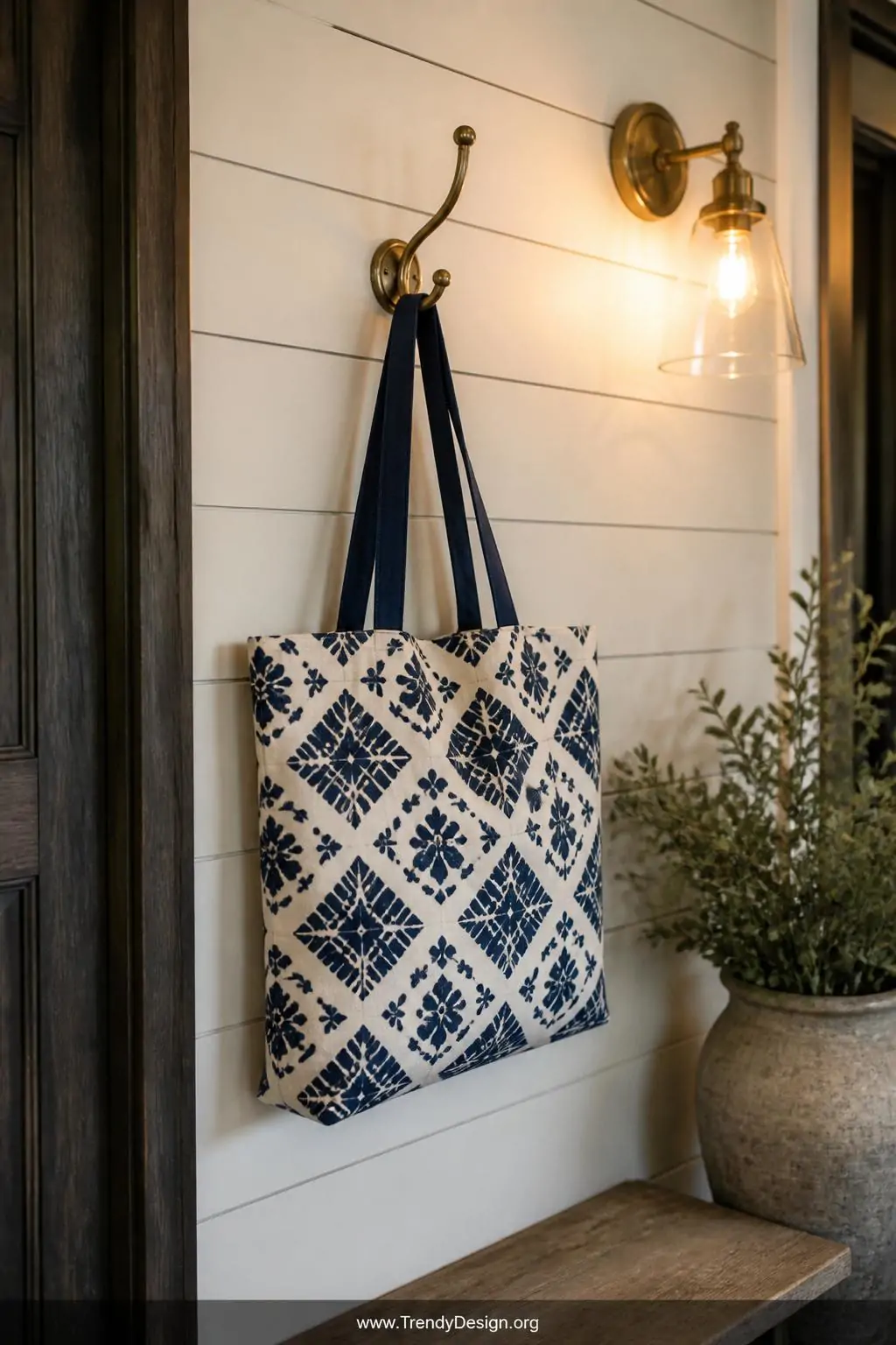 Block-printed canvas tote bag with bold geometric stamp pattern in deep indigo hanging on a brass hook against a shiplap entryway wall, warm wall sconce lighting, closeup shot of modern rustic foyer.