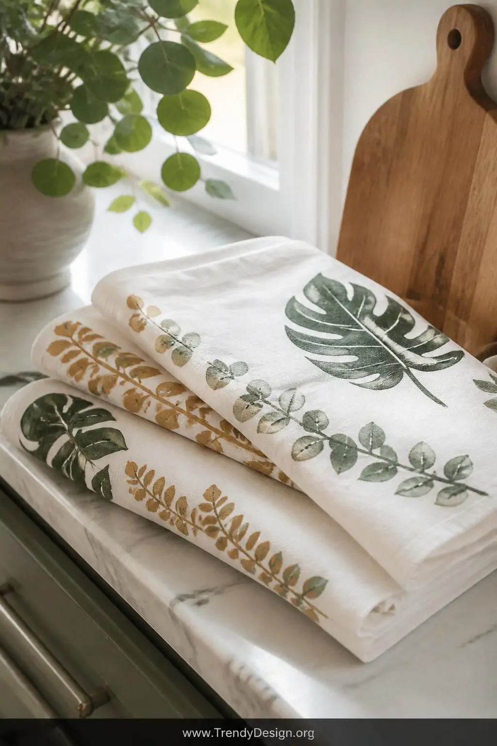 White muslin kitchen towels with eucalyptus and monstera leaf block prints in earthy green and warm ochre, folded and stacked on a marble countertop beside a wooden cutting board, bright natural window light, botanical kitchen atmosphere, closeup shot showing layered overlapping leaf border pattern