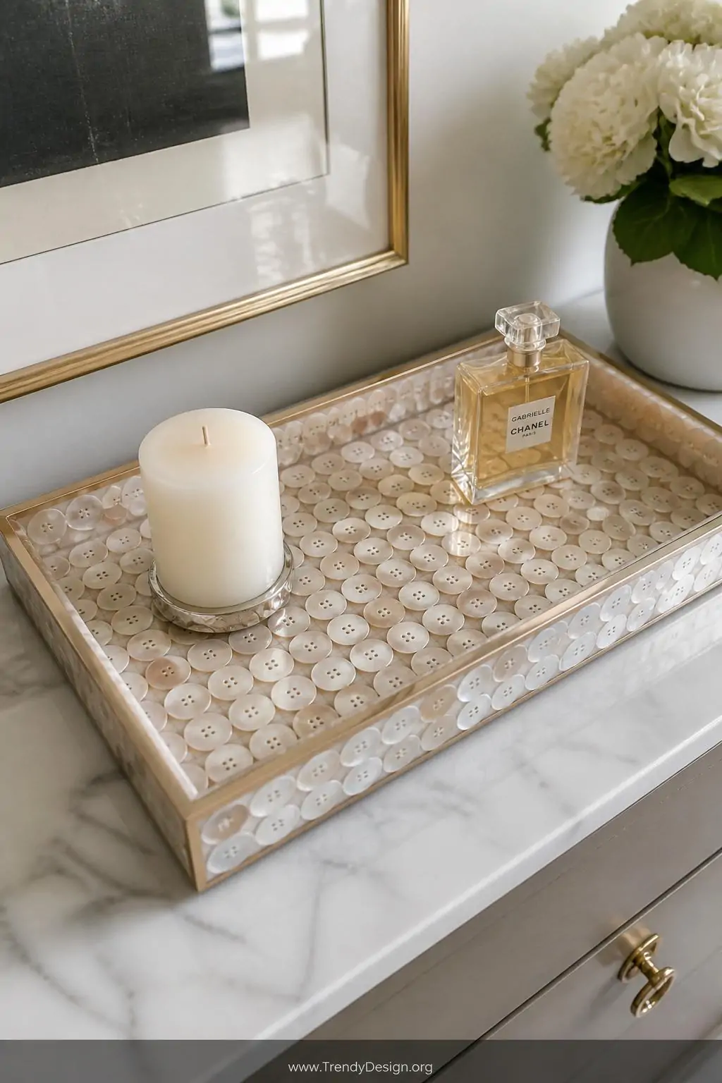 Button mosaic decorative tray on a marble console table covered in uniform shell buttons arranged in a geometric grid pattern sealed under glass-smooth clear epoxy resin, styled with a white pillar candle and perfume bottle, soft diffused natural light, medium shot from slight overhead angle.
