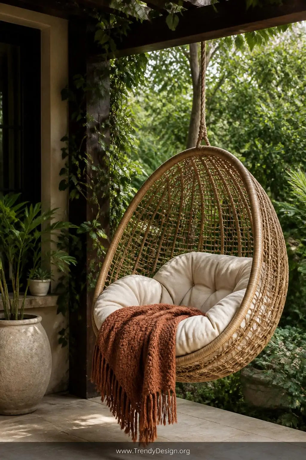 Hanging rattan egg chair with a thick cream cushion and a chunky knit rust-colored throw blanket suspended from a dark wood pergola beam in the corner of a lush green patio, soft diffused afternoon light, closeup medium shot showcasing the woven texture and dangling fringe details, photorealistic bohemian outdoor style.