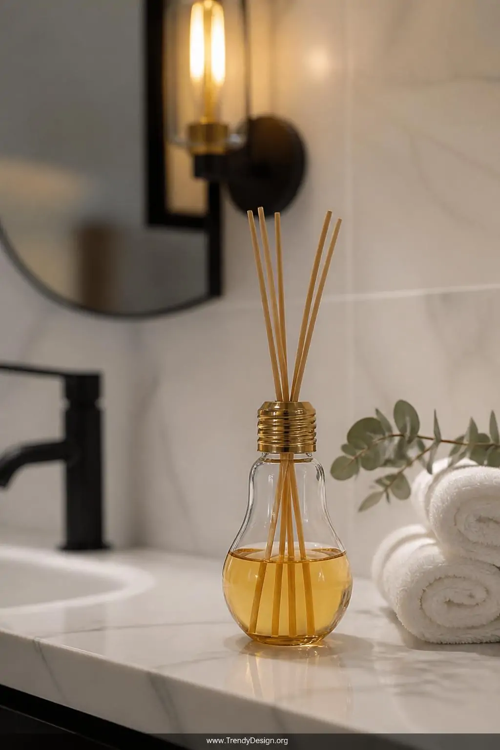 Hollowed glass light bulb reed diffuser filled with tinted amber essential oil and slender bamboo skewer reeds, sitting on a white marble bathroom vanity beside rolled cotton towels and a small eucalyptus sprig, soft warm sconce lighting, closeup shot.
