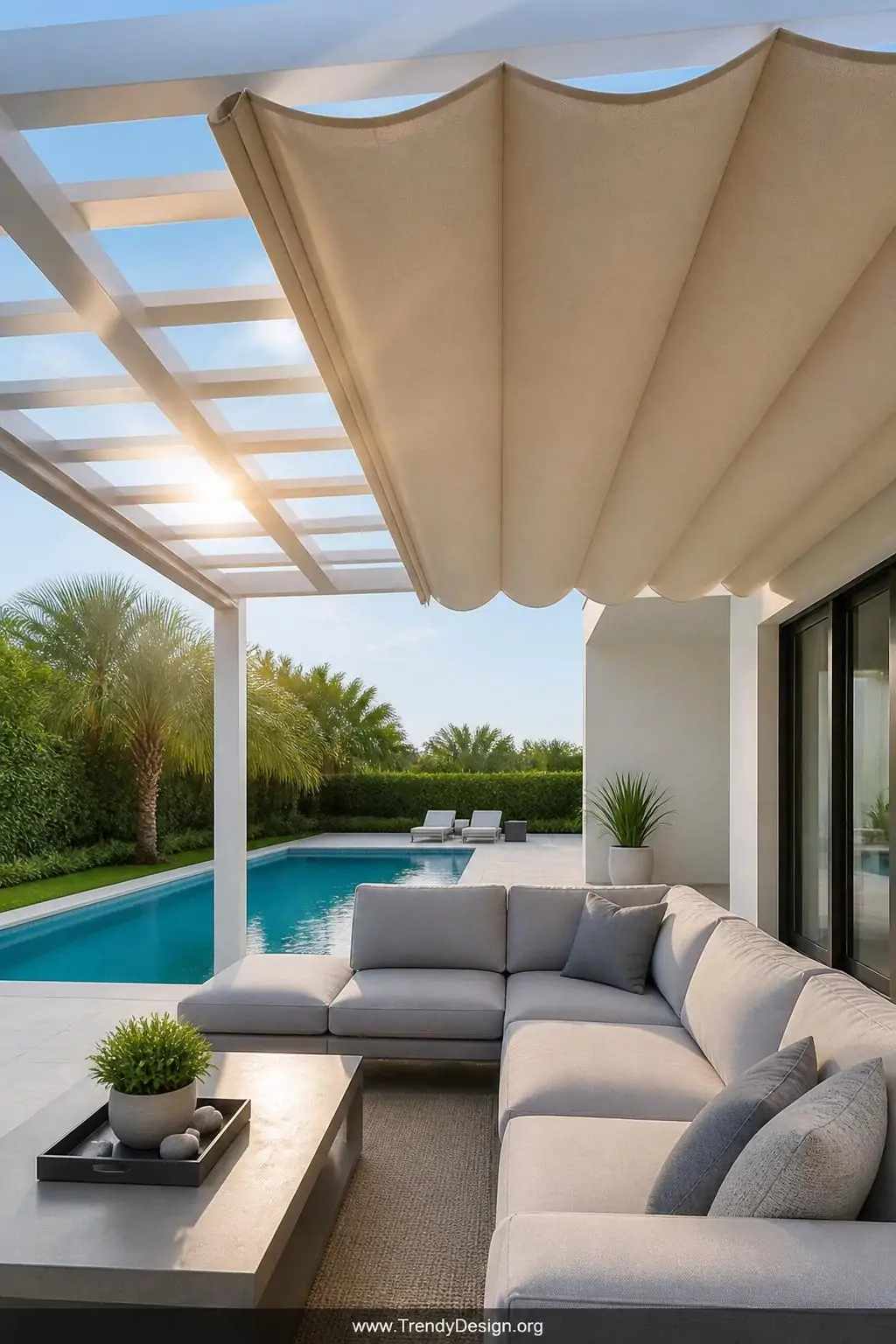Retractable fabric canopy pergola partially extended over a bright pool terrace, solution-dyed acrylic panels in warm sand tone casting soft diffused shade, clean white pergola frame visible above, sleek outdoor sectional in dove gray below, medium shot capturing the canopy mid-pull with sunlight filtering dramatically through the edge.