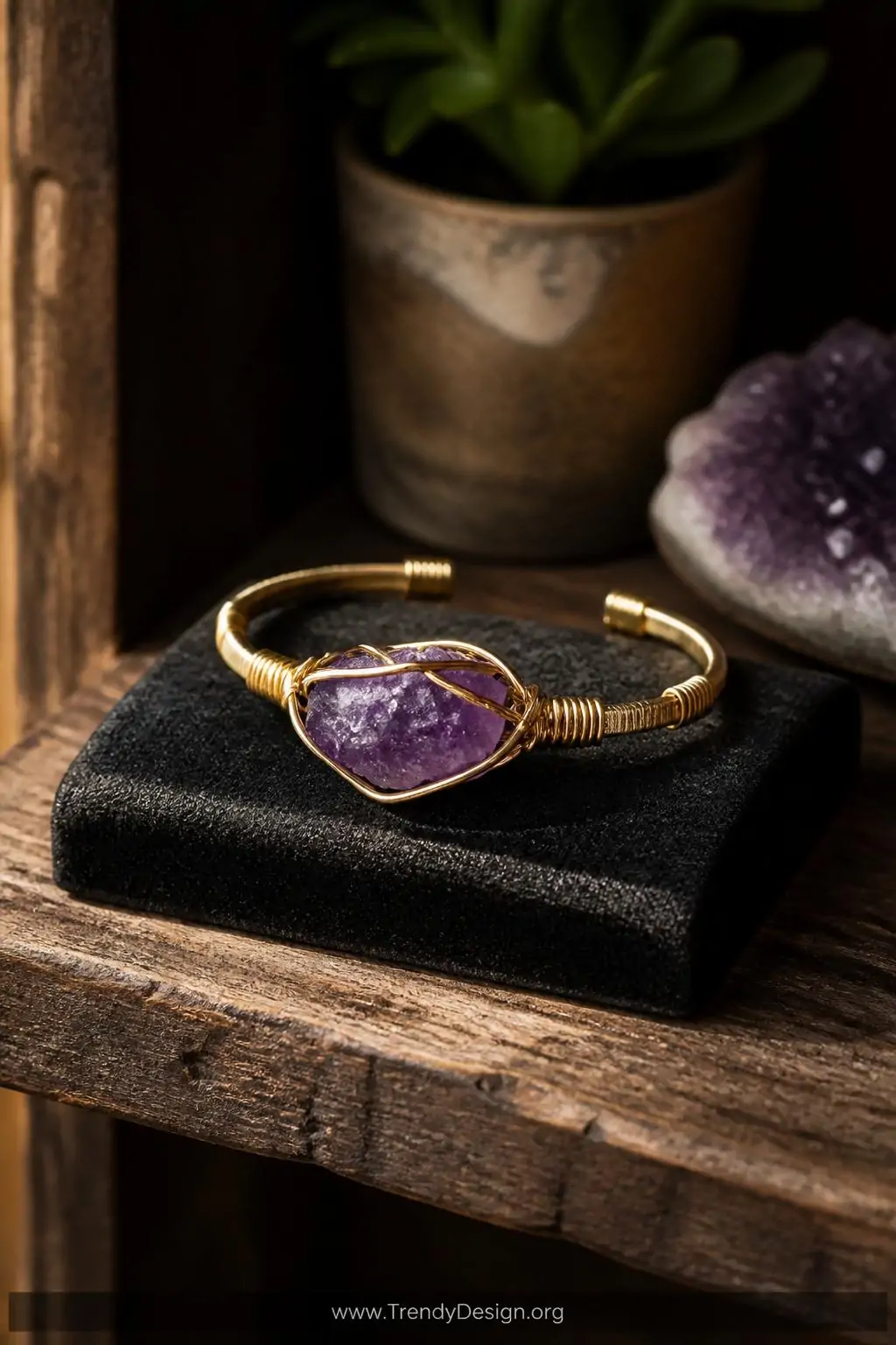 Wire-wrapped amethyst stone bracelet displayed on a dark velvet jewelry pad atop a reclaimed wood shelf, warm focused spotlight, raw purple crystal caged in delicate golden wire wrapping with natural organic edges, closeup