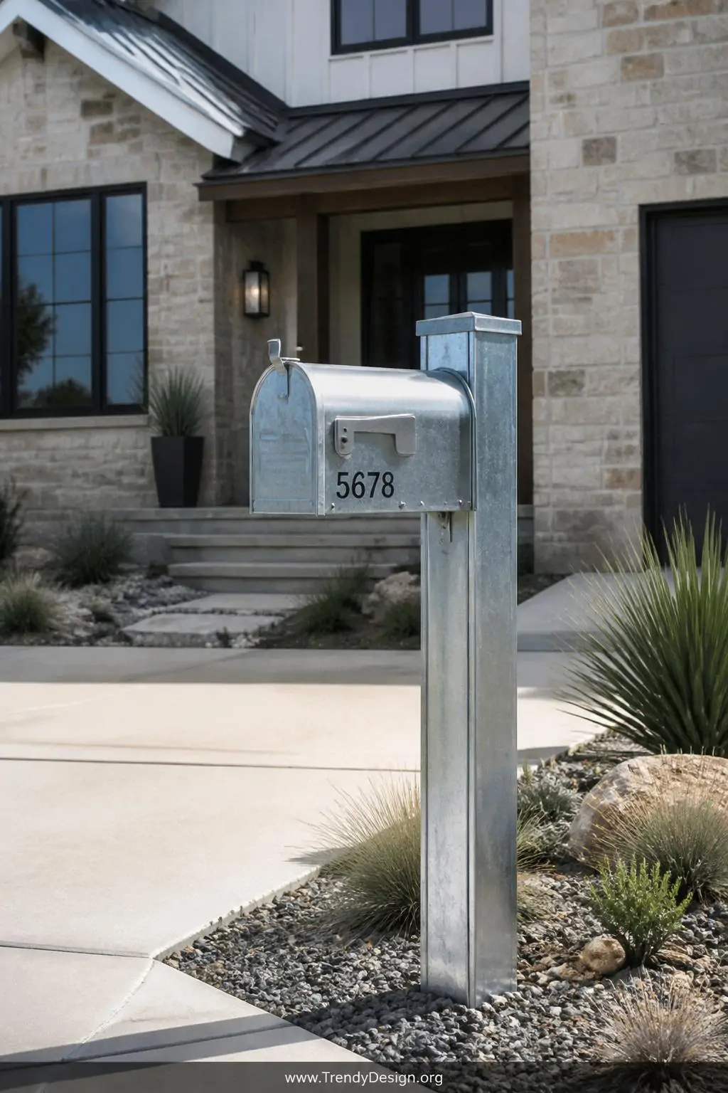 Galvanized steel mailbox post with raw zinc-coated shiny surface standing at the edge of a modern farmhouse driveway, medium shot capturing the industrial-chic reflective metallic finish against natural stone facade with drought-tolerant gravel landscaping surrounding the base.