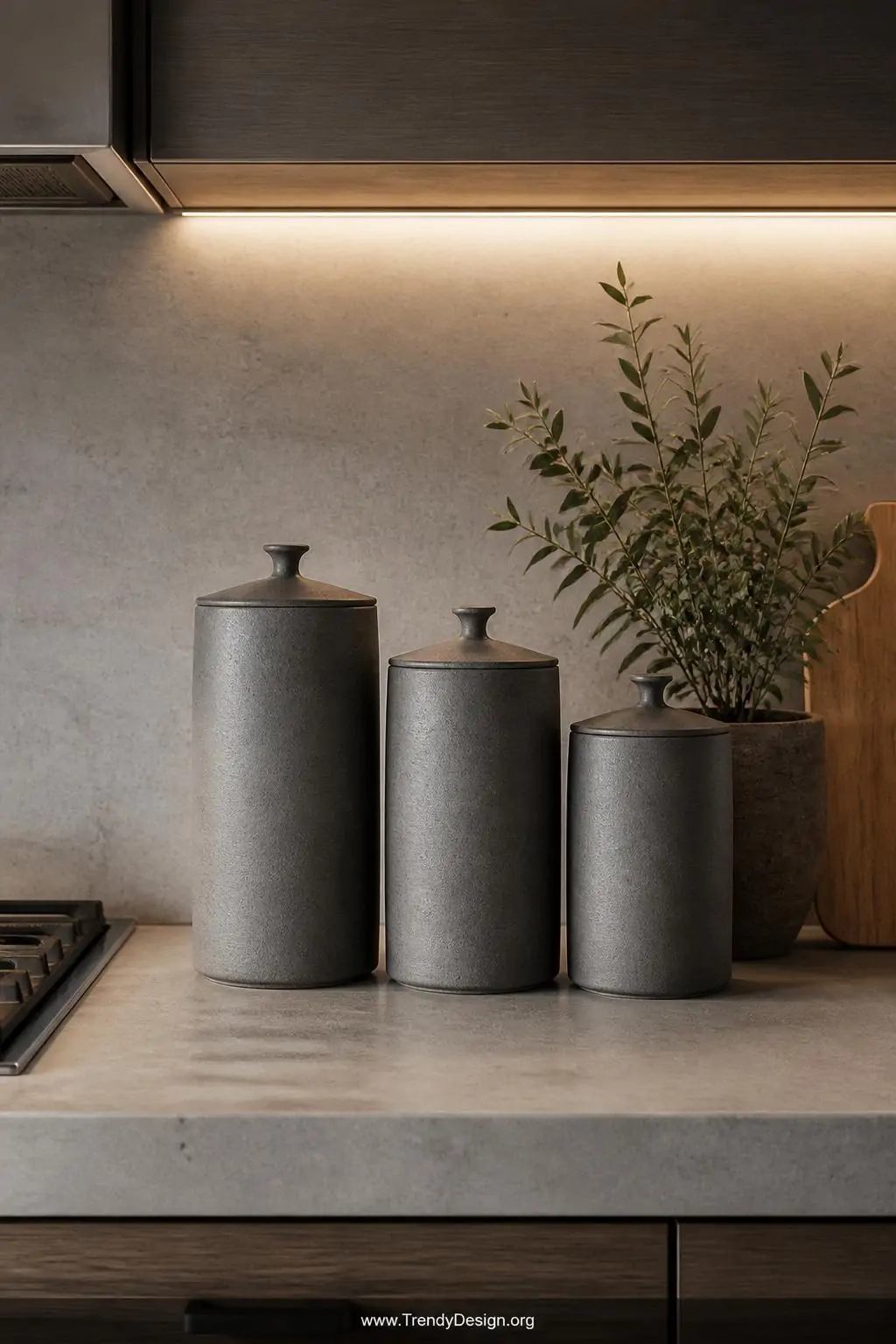 Set of three tall clay storage canisters with pinched knob lids in graduating heights sitting on a concrete kitchen counter, matte gray glaze finish, soft under-cabinet LED lighting, medium shot