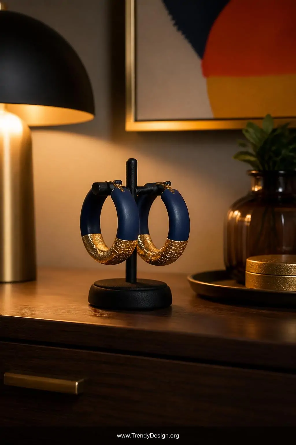 Chunky clay-wrapped hoop earrings in deep navy and gold gradient displayed upright in a small black ring stand on a dark walnut vanity table, dramatic side lamp lighting casting rich shadows, bold colorful statement pieces as the focal point, medium closeup shot.