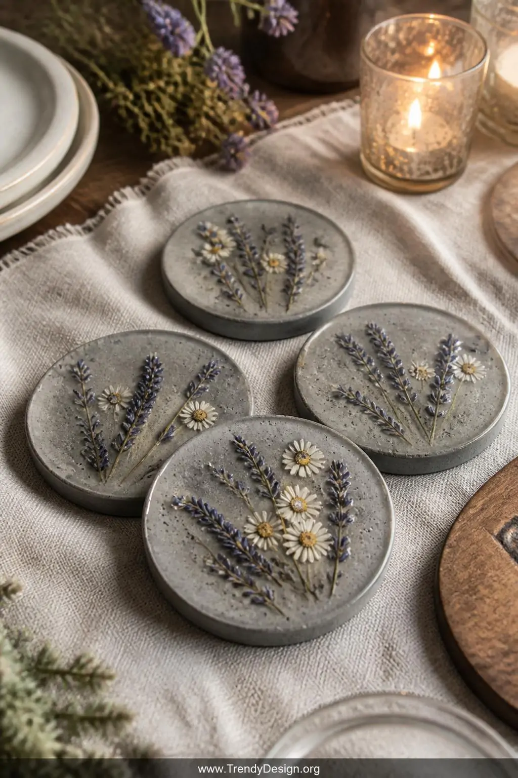 Concrete coasters with embedded dried lavender sprigs and tiny pressed daisies sealed beneath a glossy resin layer, rustic boho dining table with linen cloth and soft warm candlelight nearby, closeup shot revealing delicate botanical details preserved within the gray concrete surface.