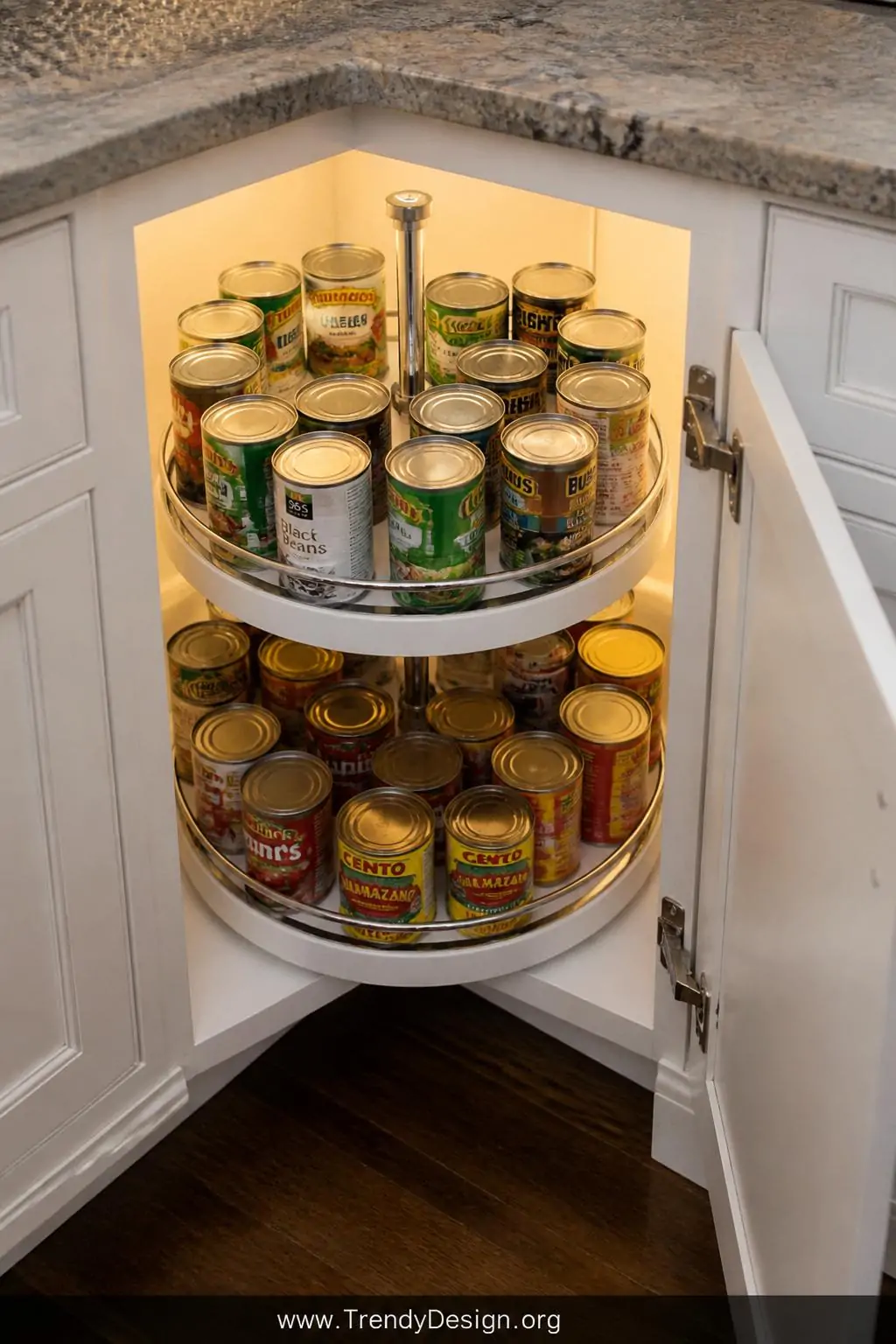 White two-tier lazy Susan turntable inside a deep corner base cabinet, traditional kitchen style, holding a full circle of mixed canned goods arranged by height with the inner and outer rings clearly visible, soft warm incandescent cabinet light illuminating the spinning chrome edge and glossy cabinet interior, medium shot looking down into the open cabinet at a slight angle.