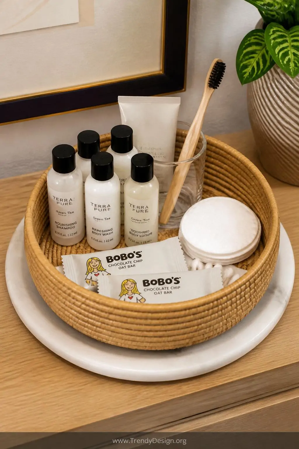 Round woven rattan basket on a white marble tray holding neatly arranged travel-sized toiletries, mini shampoo bottles, cotton rounds, a spare toothbrush, and two individually wrapped snacks on a light wood dresser top, closeup shot, photorealistic interior design.