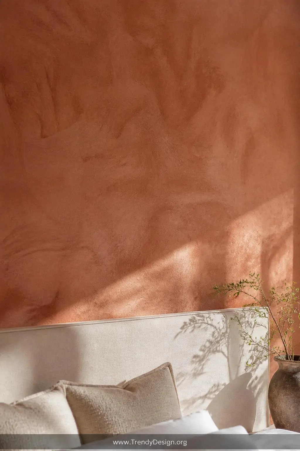 Limewash painted accent wall in warm aged terracotta with visible organic brush strokes and natural color variation catching directional afternoon light, closeup detail shot of the textured plaster-like surface above a simple cream headboard where the shifting depth and movement of the finish creates an artisan European villa quality.