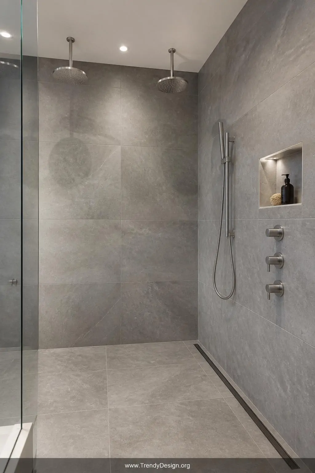 Dual overhead rainfall showerhead and sleek brushed chrome handheld fixture above a continuous linear floor drain running flush along one wall beside a matching recessed niche in a contemporary walk-in shower, uninterrupted large-format grey tile flowing seamlessly across floor and walls, medium shot, photorealistic interior design.