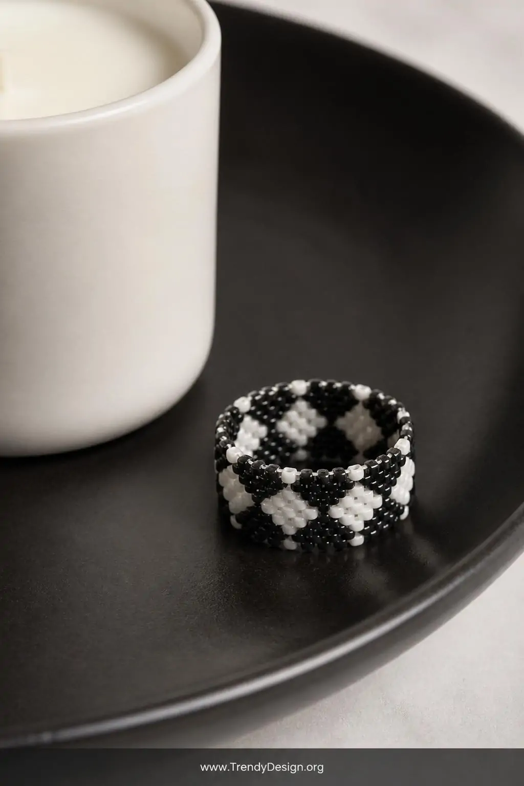 Geometric black and white harlequin diamond pattern seed bead ring photographed flat on a matte black ceramic dish beside a minimalist white candle, crisp studio-style directional light, closeup revealing sharp angular stitch precision and graphic contrast