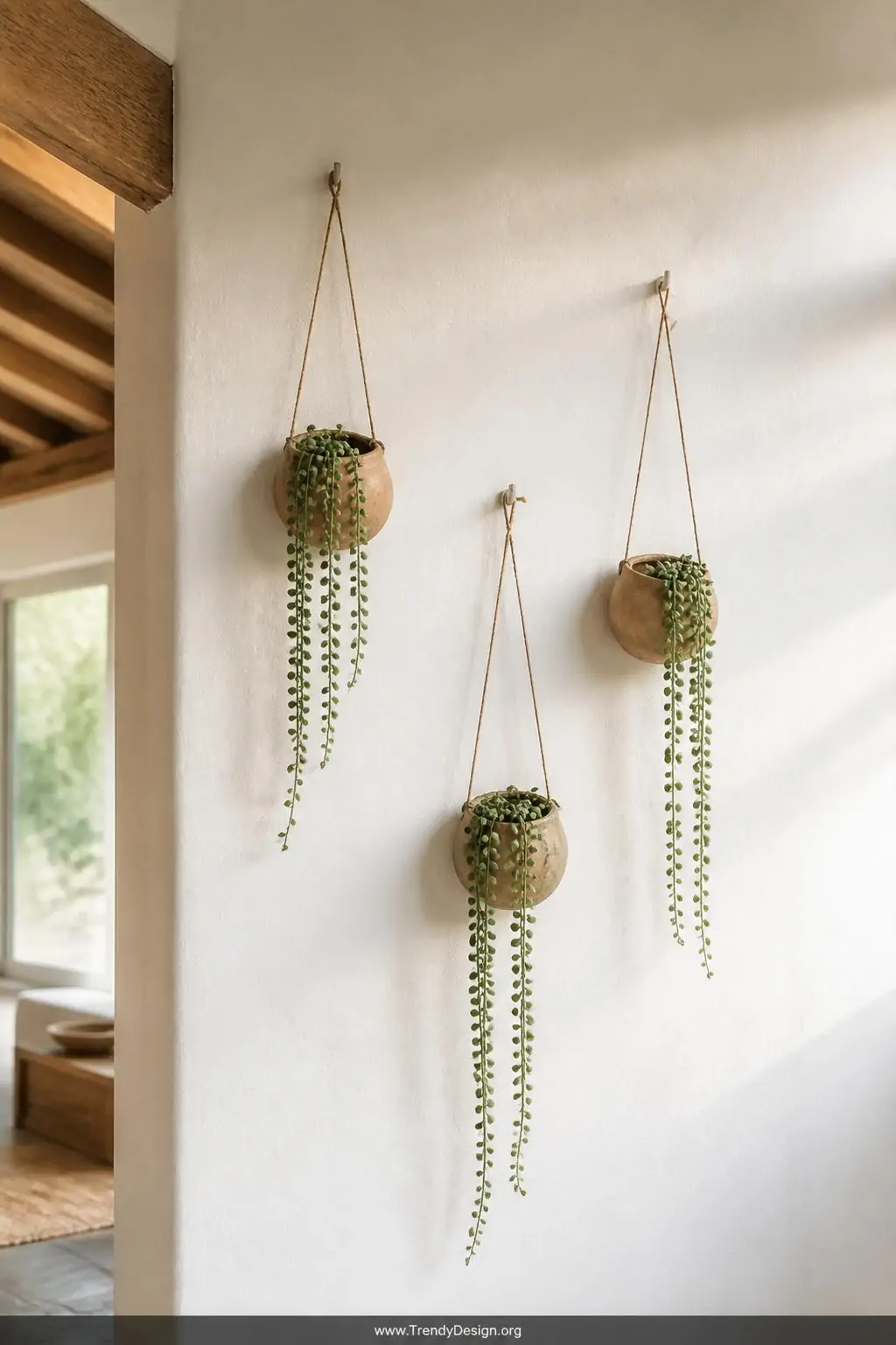 Three small clay pinch pot wall planters in earthy tones hung at varying heights with jute twine on a white plaster wall, trailing string of pearls cascading from each pot, natural side light, wide shot