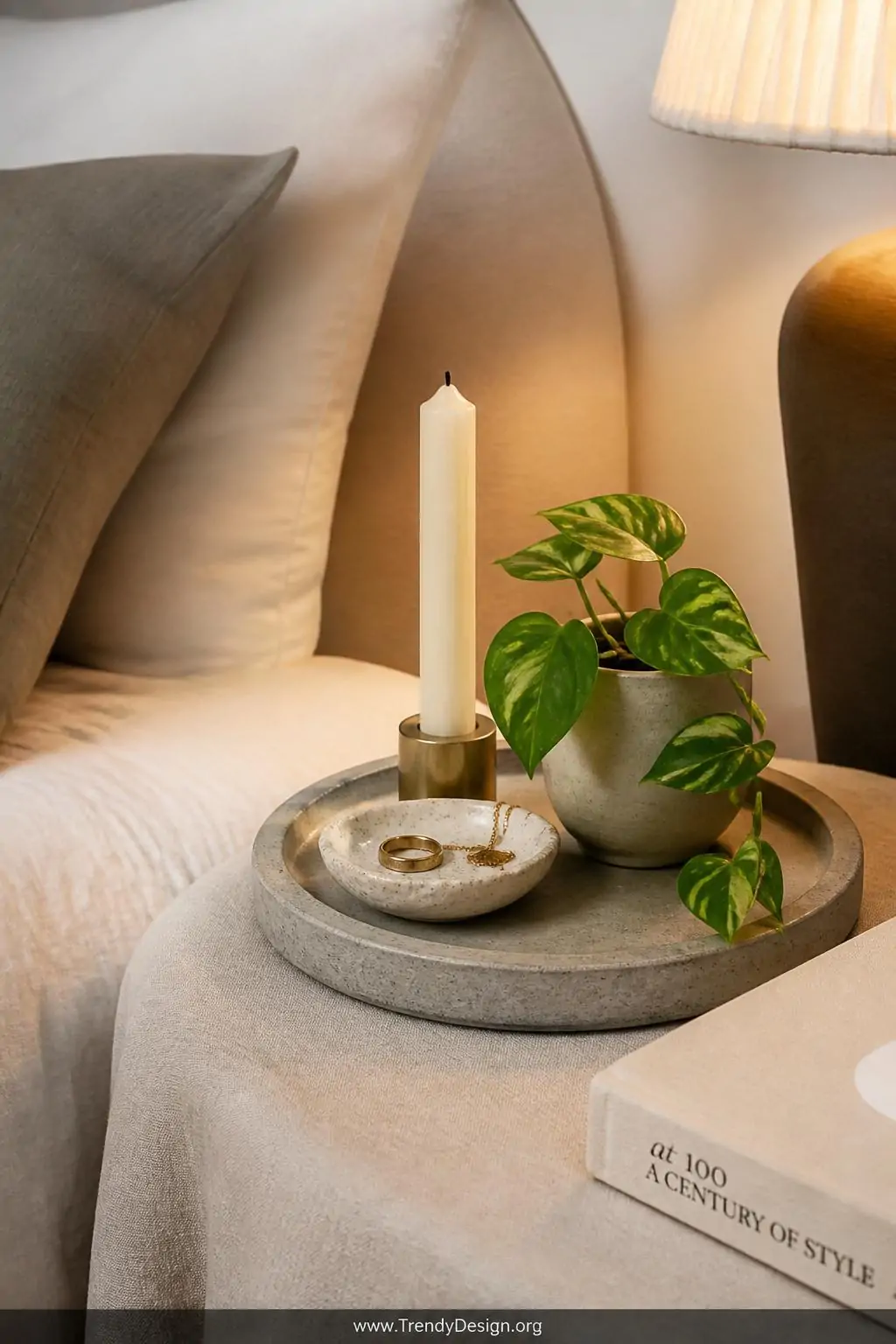 Small round concrete tray on a linen-draped nightstand in a soft neutral bedroom, styled with a cream taper candle, a delicate ring dish holding gold jewelry, a tiny trailing pothos plant, and a hardcover book, warm bedside lamp glow, closeup shot.