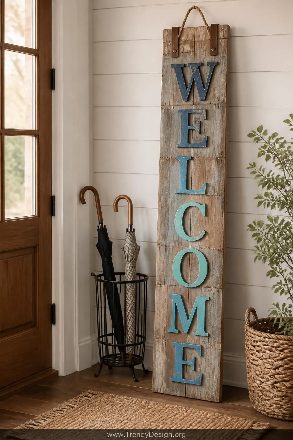 Tall reclaimed pallet wood welcome plank sign with vertical lettering in graduated dusty blue tones, leaning against a shiplap entryway wall beside a woven jute rug and black iron umbrella stand, a leather rope hanger at the top, wide shot with warm afternoon light.