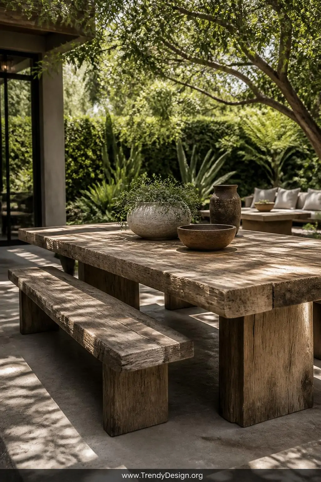 Thick outdoor teak wood slab bench-table combo on a garden patio surrounded by greenery, rustic outdoor living style, wide shot, dappled natural sunlight filtering through trees and casting organic shadows across the chunky weathered slab surface and sturdy leg joints, photorealistic interior design, no people