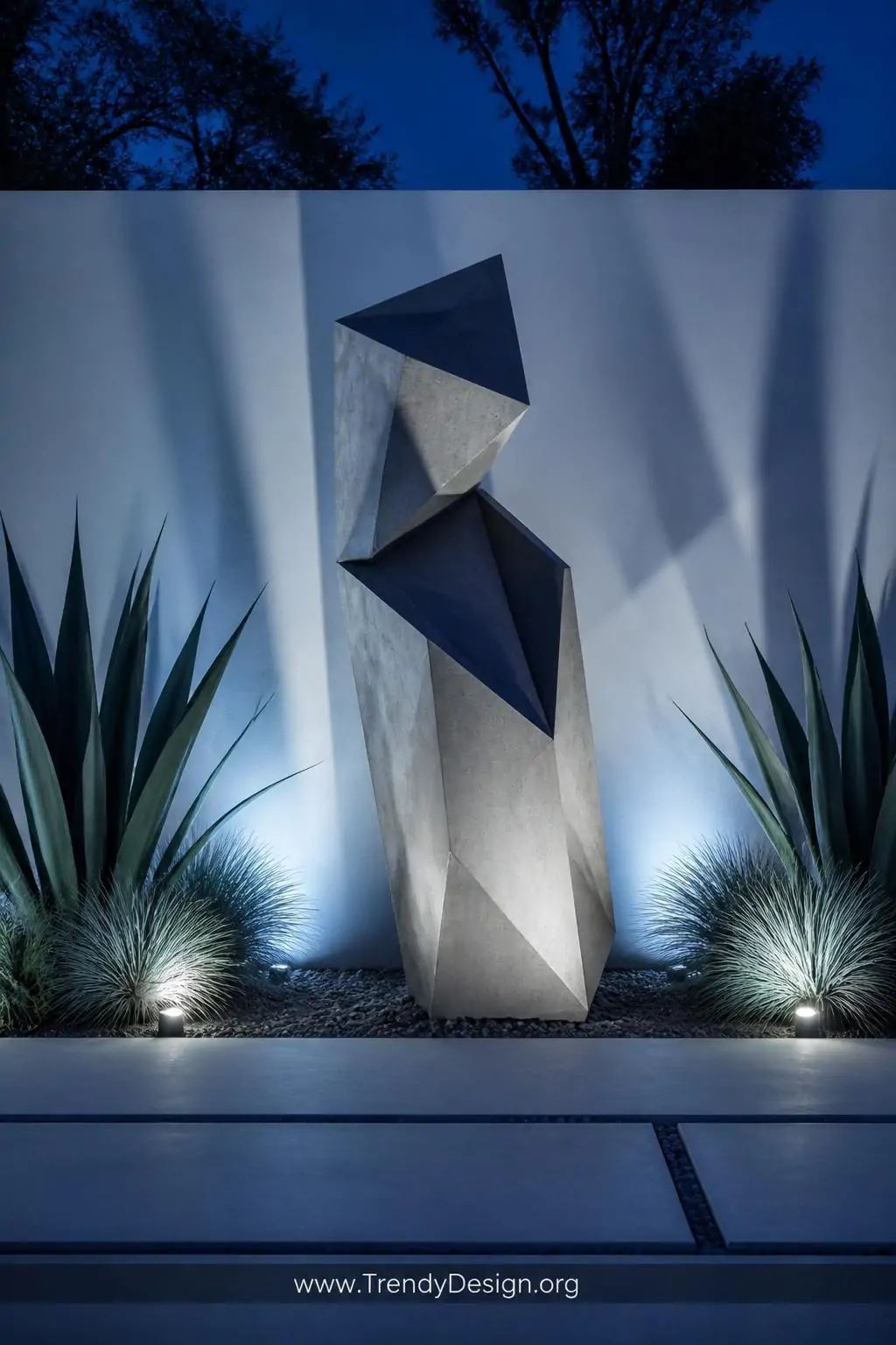 A bold geometric abstract concrete sculpture stands alone against a clean white rendered garden wall, its smooth angular plan