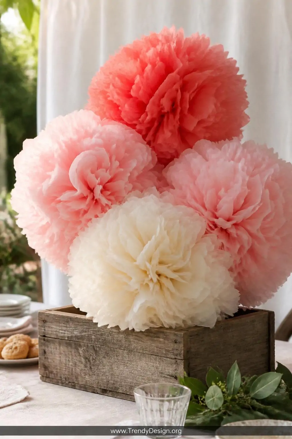 Cluster of four oversized fluffy tissue paper pom-poms in coral, blush, and cream nestled at varying heights inside a low weathered wooden box on a garden party table, soft afternoon natural light, medium shot capturing the voluminous layered texture against a pale linen backdrop.