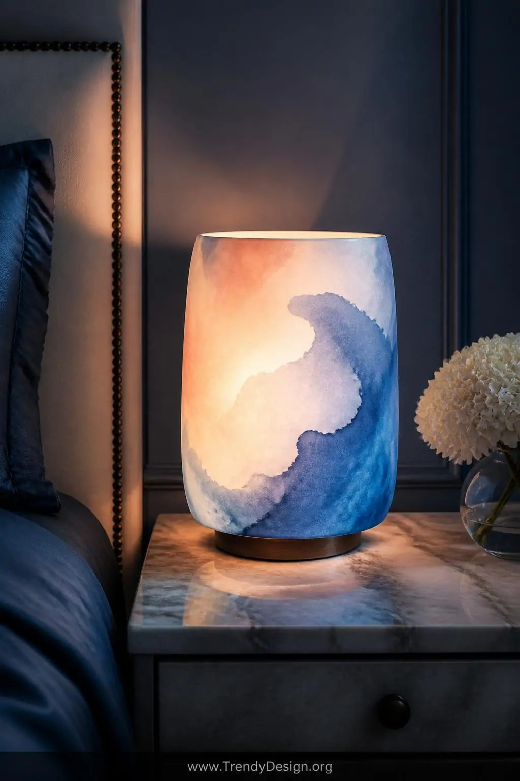 Watercolor-effect fabric lampshade painted in translucent blues and soft peach casting dreamy colored light across a marble nightstand, glowing from within against a dark moody bedroom wall, closeup shot of contemporary bedroom.