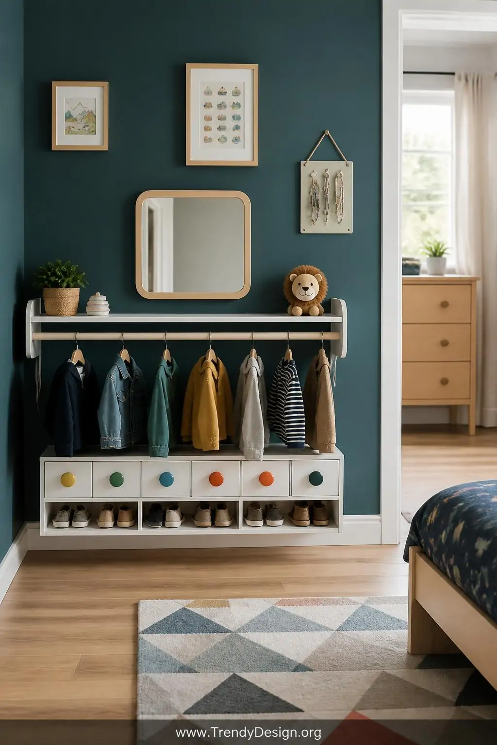 Low white wooden clothing rod with colorful drawer pulls and open shoe cubby mounted against a deep teal accent wall with a small mirror at child height, medium shot of a Montessori dressing station in a child's bedroom, bright even morning light