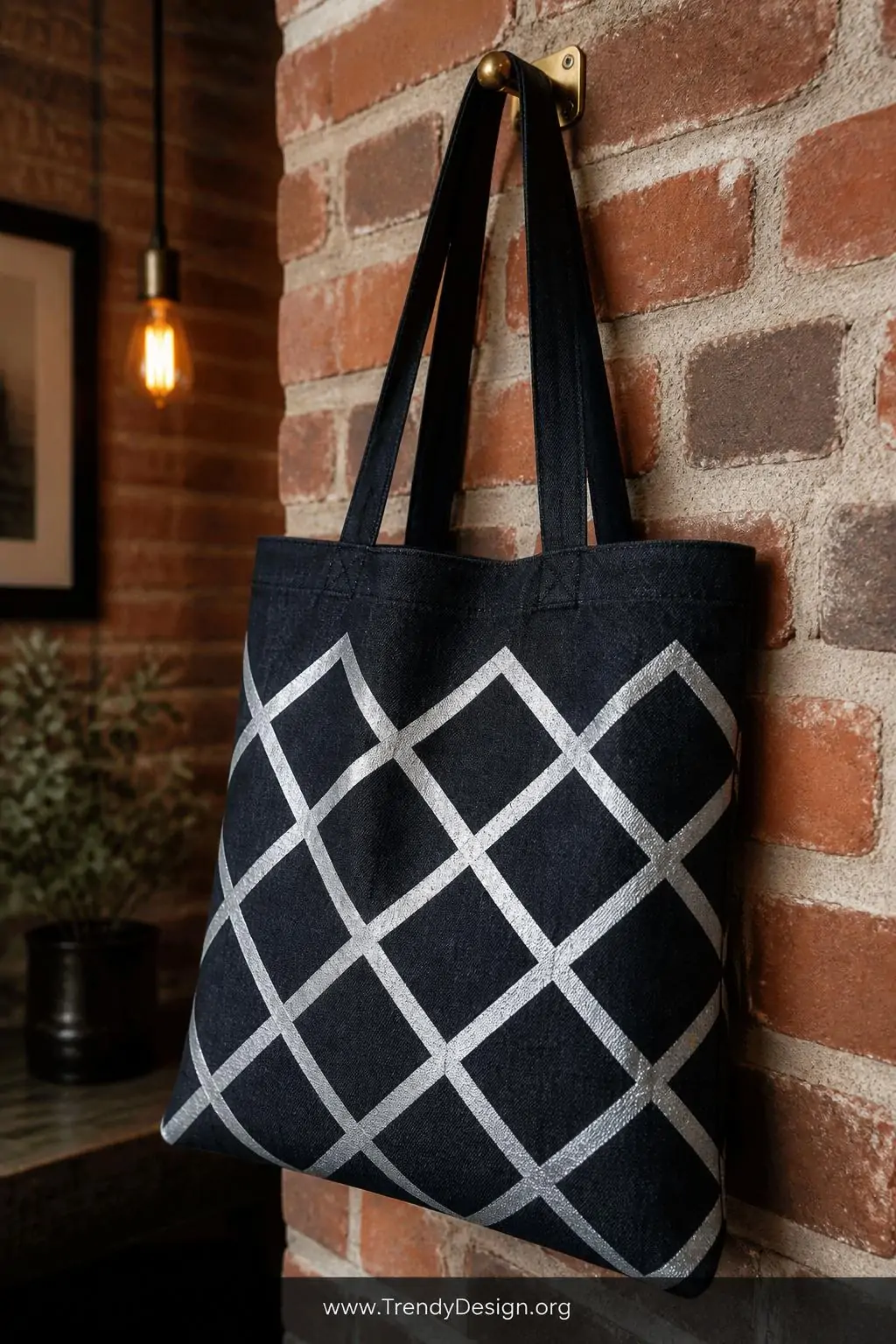 Black denim tote bag with bold white metallic diamond grid block-printed pattern hanging on a brass wall hook, exposed brick wall, warm Edison bulb lighting, crisp geometric lines popping against dark structured denim weave, medium closeup shot