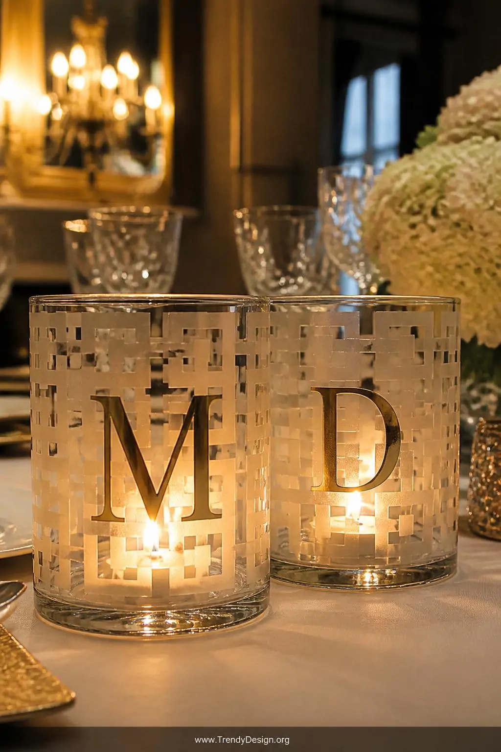 Clear glass candle holders with frosted geometric vinyl cutouts and metallic gold monograms glowing on a dining table, candlelight flickering from inside illuminating the etched silhouettes, closeup showing the luminous contrast between warm flame and frosted vinyl.