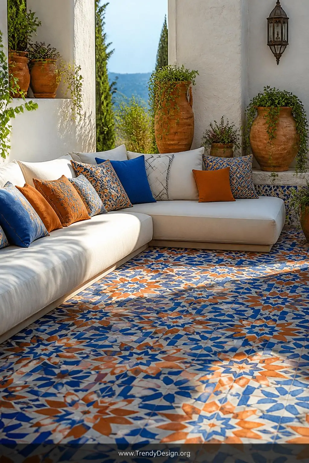 Moroccan tile-print outdoor rug in vibrant cobalt blue, burnt orange, and white beneath a low whitewashed sectional sofa, medium shot, terracotta pots bursting with trailing greenery, bright Mediterranean afternoon light illuminating the repeating star and cross geometric pattern against sun-bleached walls.
