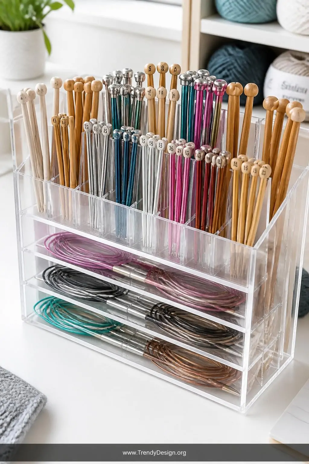 Clear acrylic divided organizer on a white craft desk displaying straight knitting needles stood upright in narrow compartments and circular needles laid flat in wider sections, cool bright overhead lighting, transparent walls showing every needle clearly, crisp organized grid layout, closeup shot