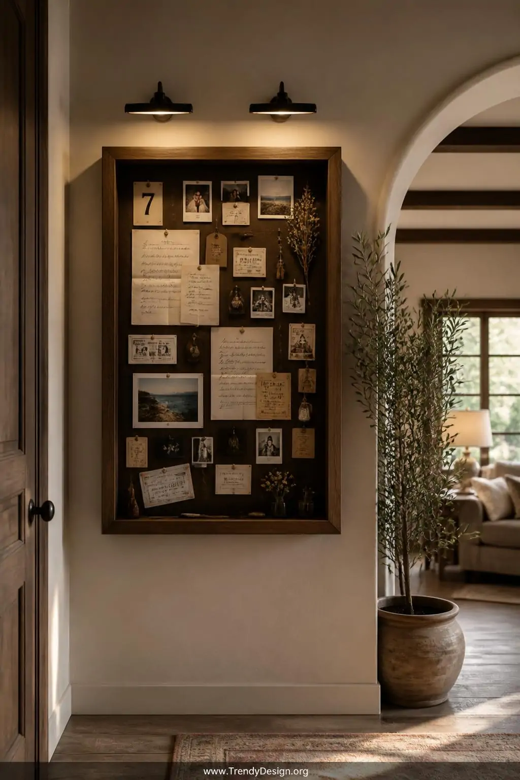 Deep wooden shadow box frame mounted on an entryway wall displaying folded notes, ticket stubs, tiny photographs, and small meaningful objects arranged in a living collage, warm gallery wall lighting from overhead fixtures, wide shot showing surrounding neutral plaster wall and adjacent greenery, photorealistic interior design.