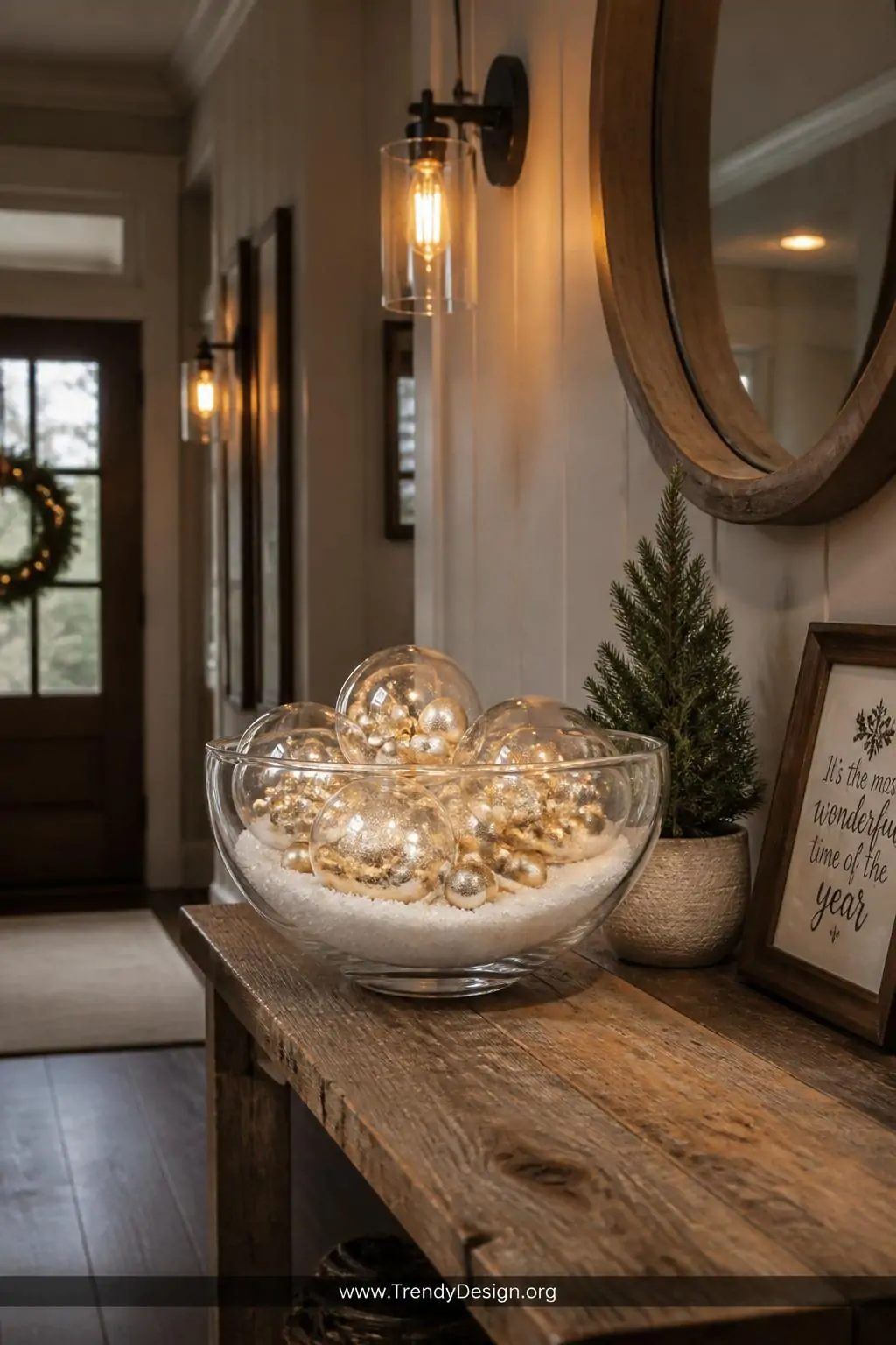Glass bowl centerpiece filled with clear balloons stuffed with miniature gold ornaments and faux white snow on a rustic reclaimed wood entryway table, warm Edison bulb sconce lighting casting a festive holiday glow, medium shot