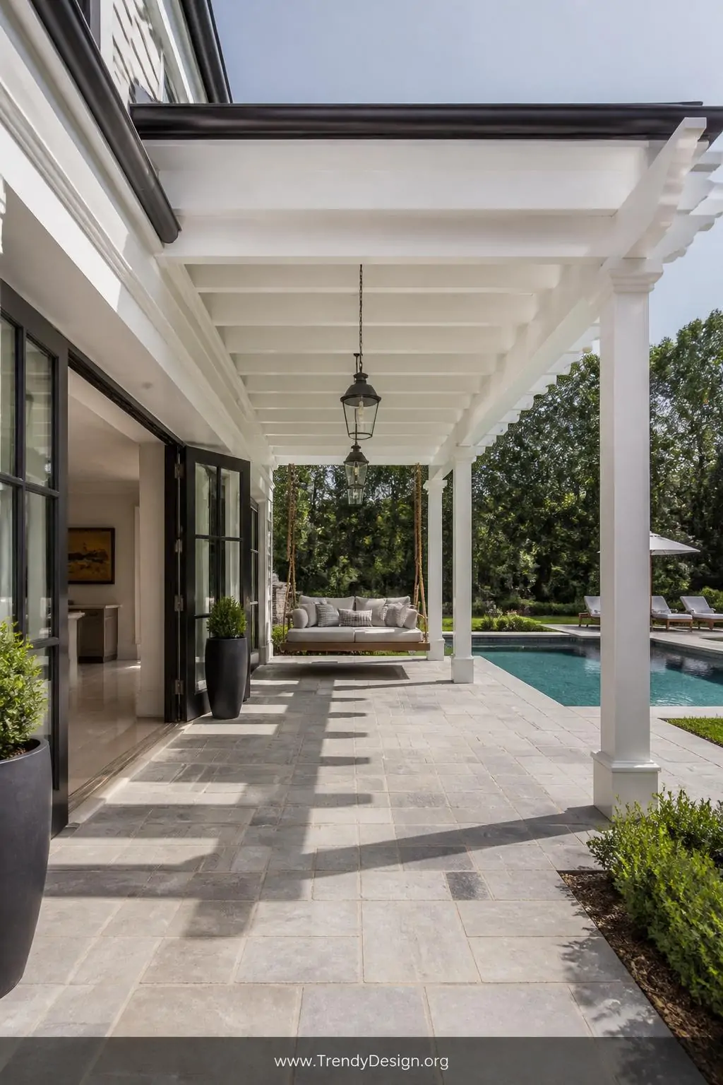Attached pergola extending directly from French doors over a pool patio, matching the home's exterior in crisp white with dark trim, pendant lights hanging from the beams, flowing seamlessly from indoor tile to outdoor stone pavers, a hanging daybed swing visible in the shade, wide architectural shot showing the home-to-pergola connection.