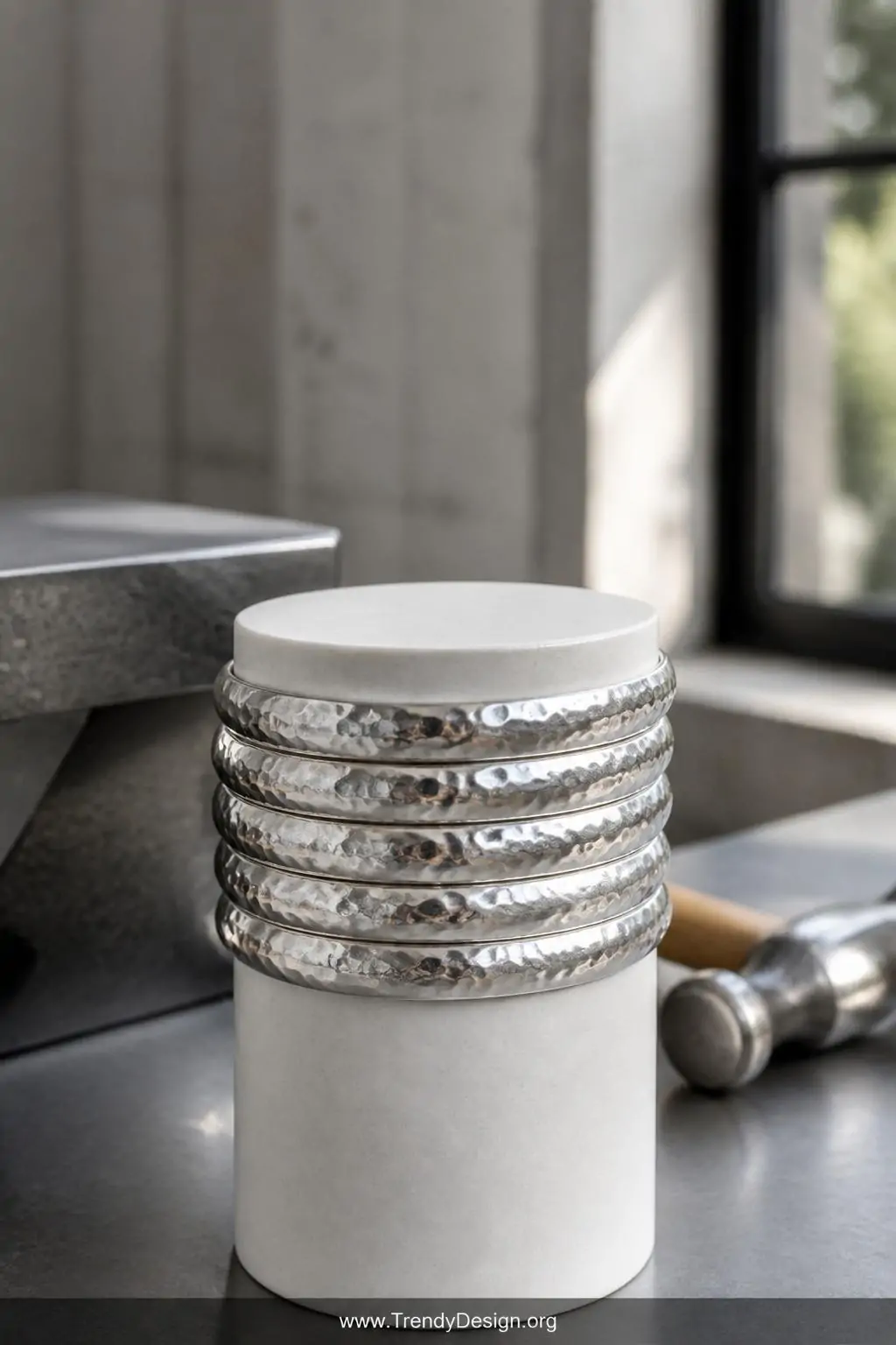Hammered sterling silver bangles stacked six high on a cylindrical white ceramic stand, bright directional light creating multi-directional reflections across dimpled metallic surfaces, steel bench block and ball-peen hammer blurred in background, wide shot