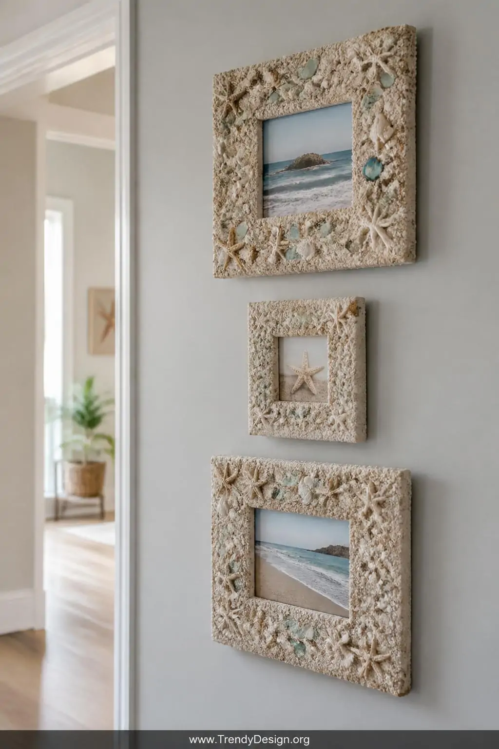 Gallery wall arrangement of three sand and shell-encrusted picture frames on a pale gray hallway wall, each frame densely textured with fine sand and pressed tiny starfish and sea glass, bright natural sidelight, medium shot