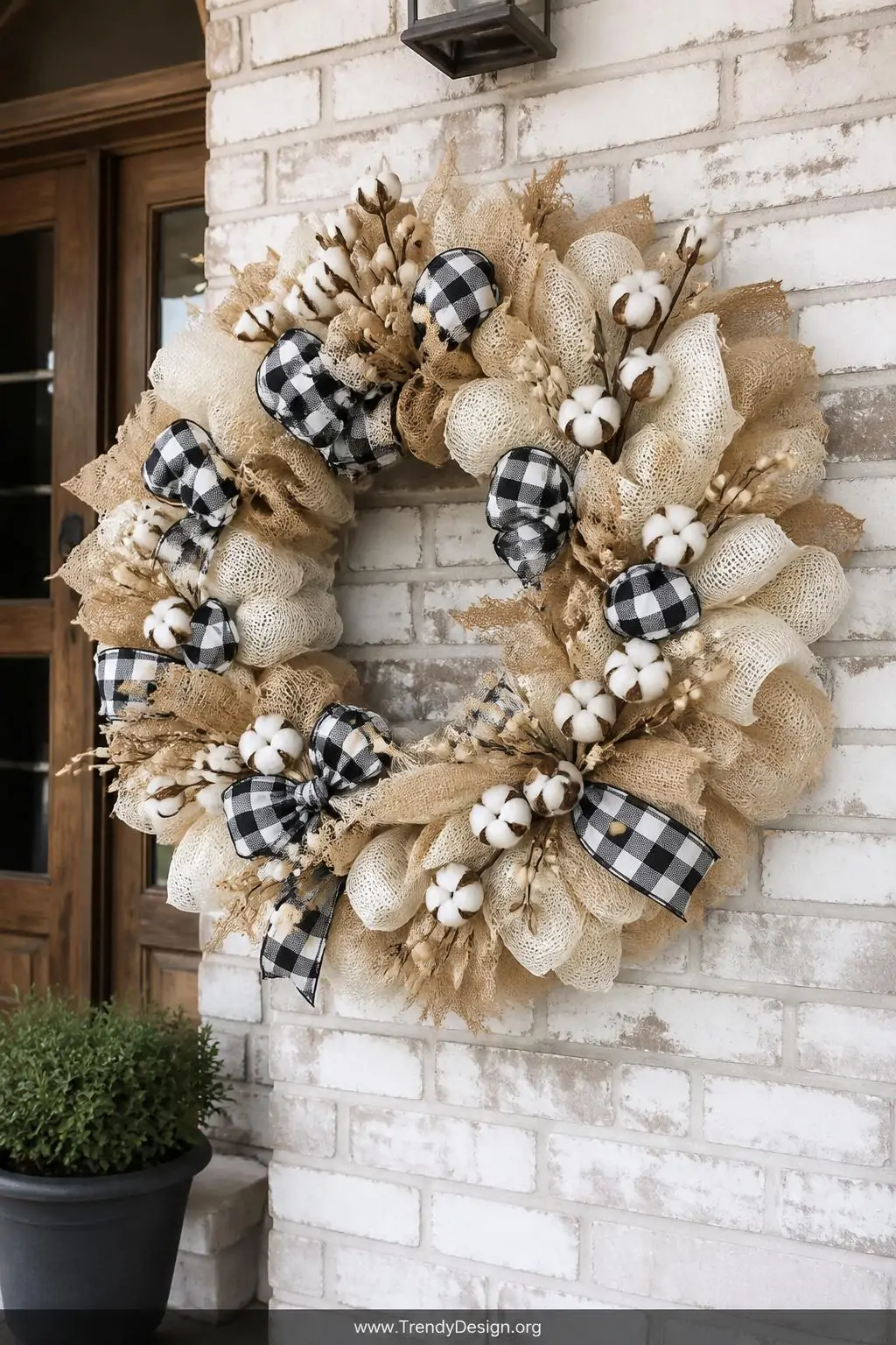 Cream and tan deco mesh wreath featuring bold black and white buffalo check ribbon, cotton stems, burlap accents, and wooden bead picks resting against a whitewashed brick entryway wall, diffused neutral daylight, wide shot.
