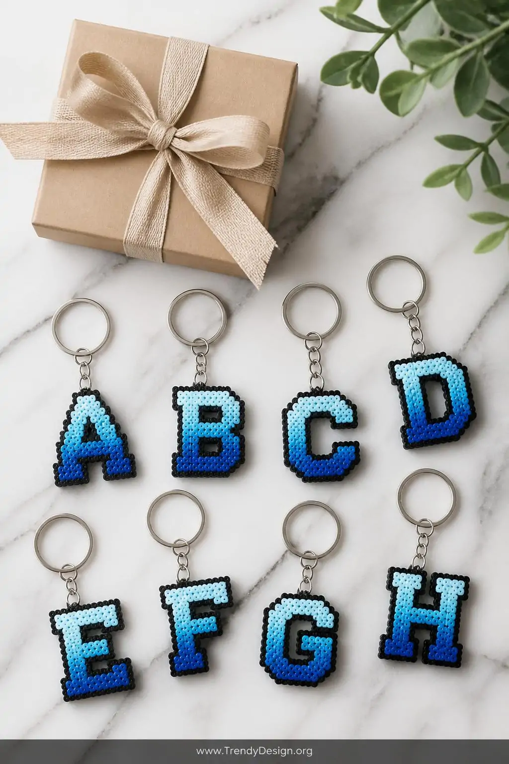 Block letter initial Perler bead keychains in gradient blue tones arranged on a marble surface beside a gift box with kraft paper ribbon, clean minimalist styling, bright diffused light, medium flat lay shot